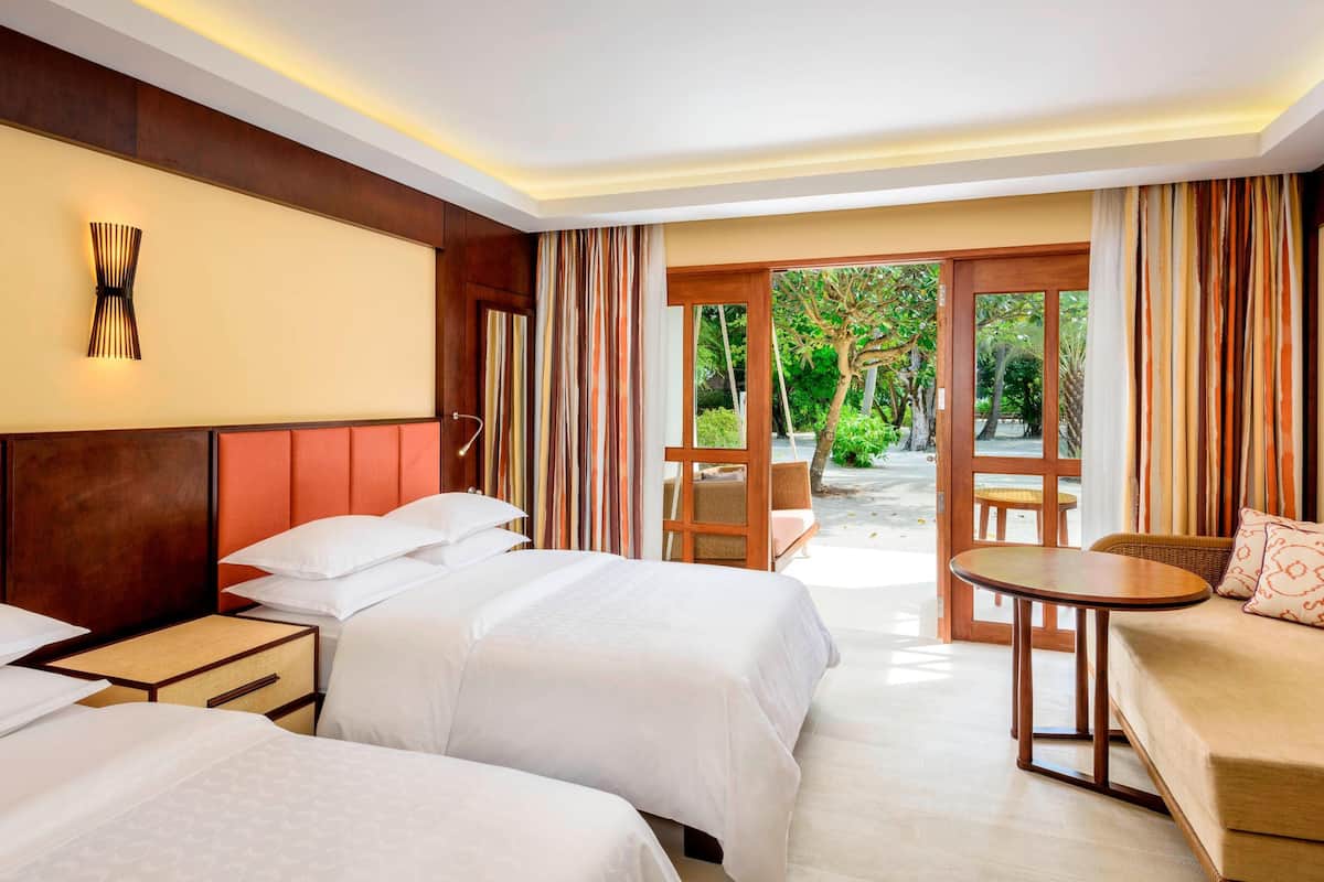 A hotel room with two beds, a sofa, a small table, and a view of a garden through a glass door.