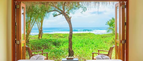A room with two beds, a small table, and chairs, overlooking a beach with palm trees and the ocean.