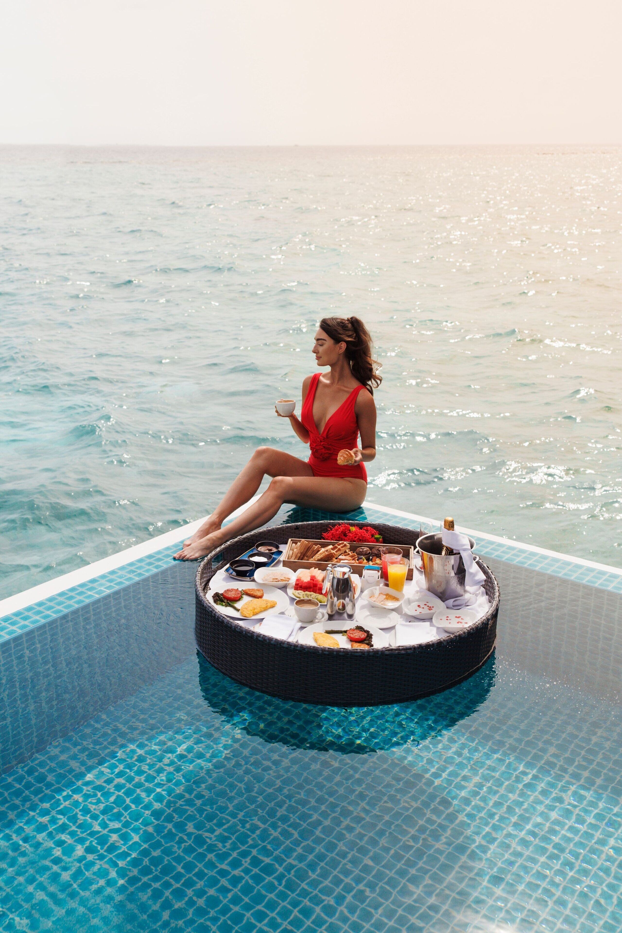 Una mujer con traje de baño rojo sentada en una plataforma flotante junto a una piscina, con una bandeja de desayuno.