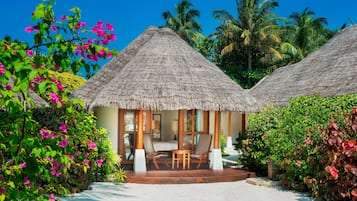 A thatched-roof bungalow with a wooden deck, surrounded by lush greenery and palm trees.
