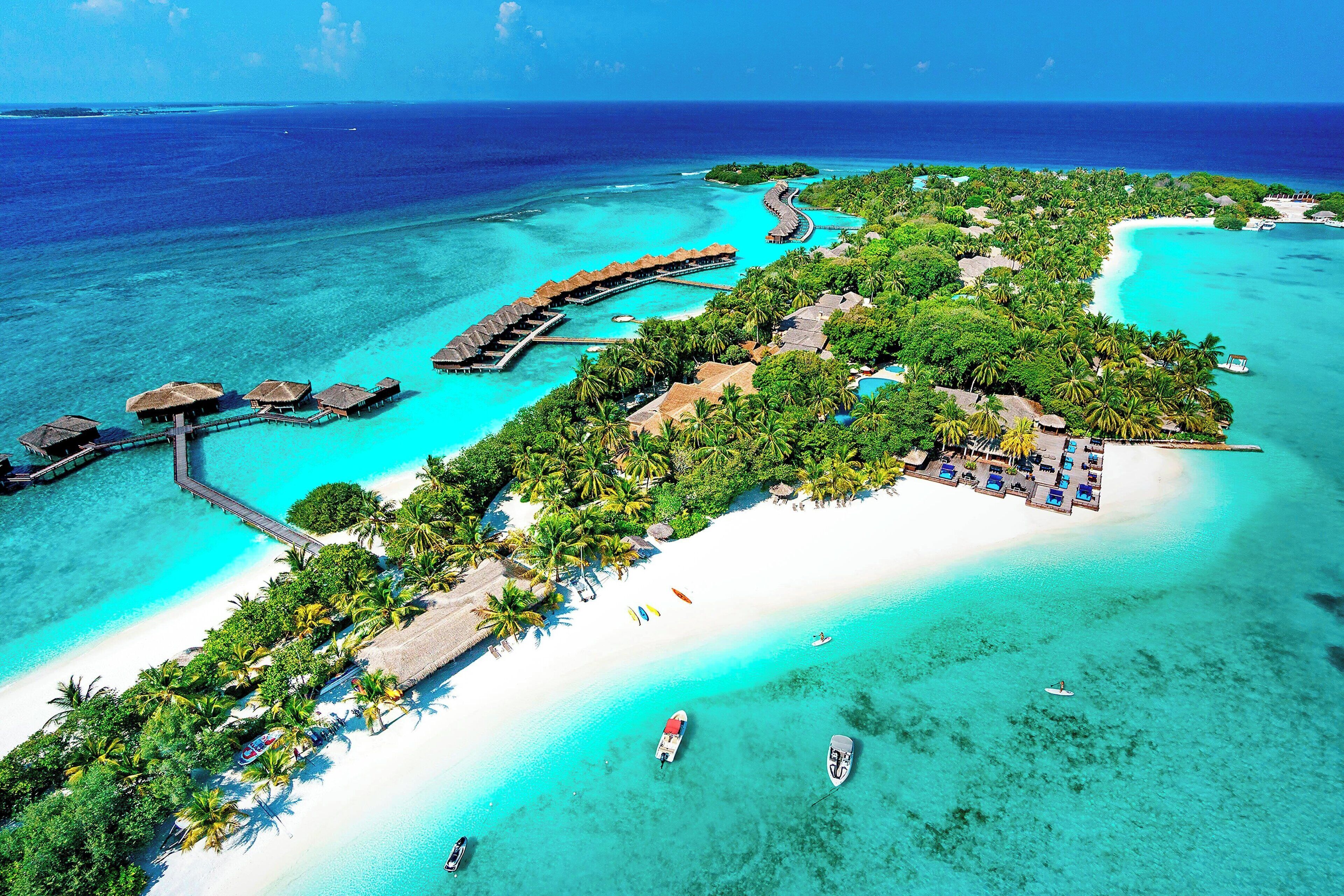 An aerial view of a tropical resort with overwater bungalows, a white sandy beach, and clear turquoise waters.