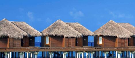 Overwater bungalows with thatched roofs, wooden exteriors, and a clear blue sky.