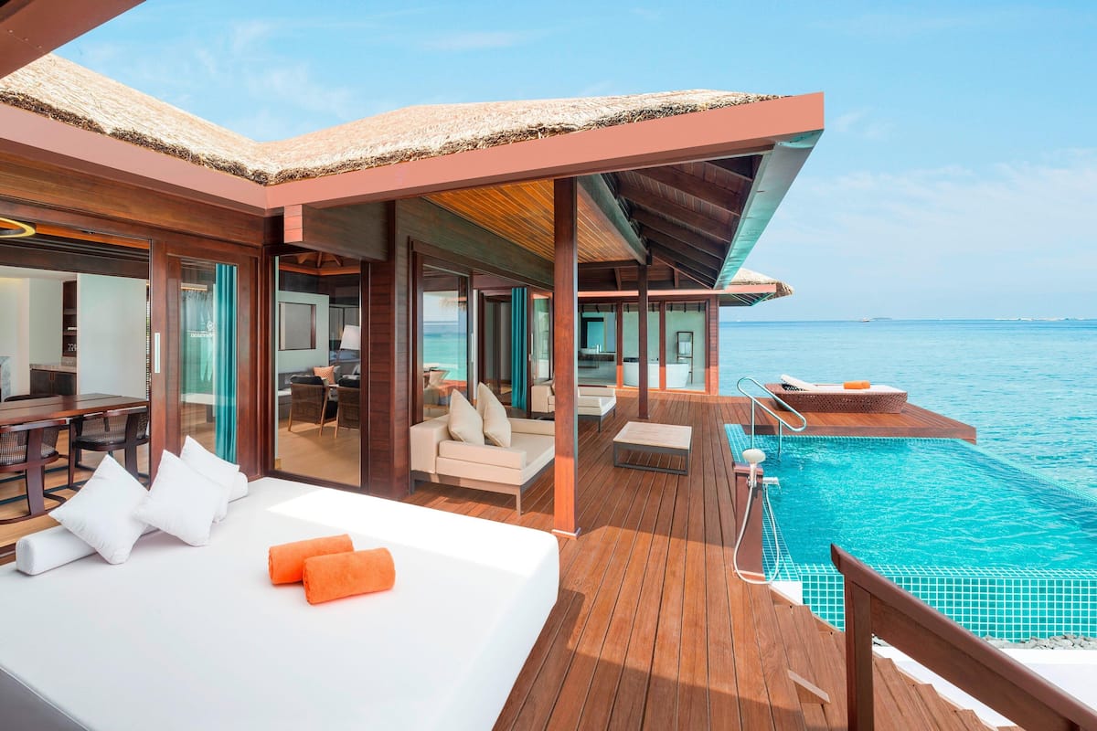 A luxurious overwater villa with a private pool, wooden deck, and a cozy sitting area with a sofa and chairs.