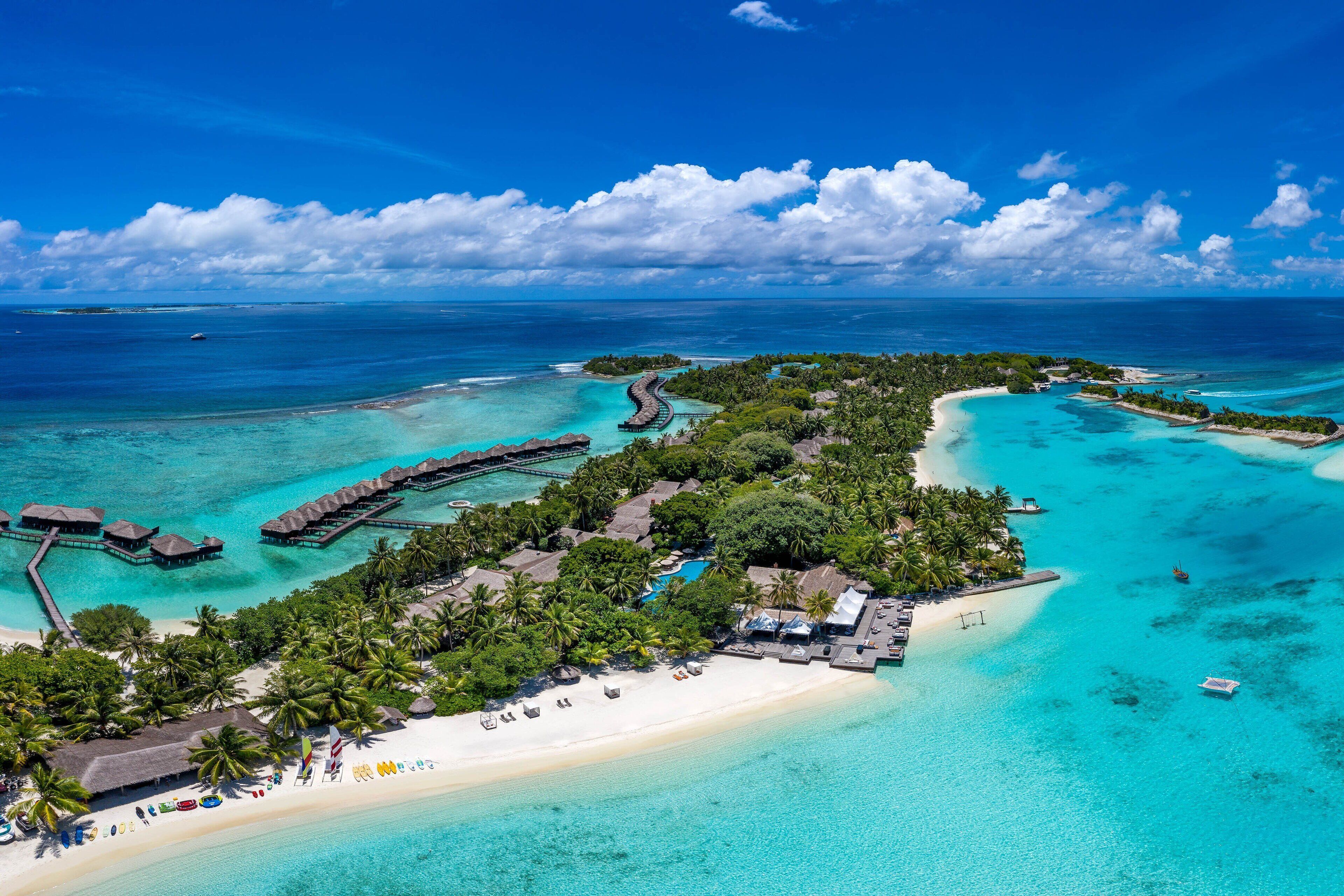 An aerial view of a tropical resort with overwater bungalows, a sandy beach, and clear turquoise waters.