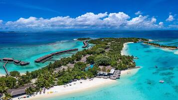 Vue aérienne d’un complexe hôtelier tropical doté de bungalows sur pilotis, d’une plage de sable et d’eaux turquoise cristallines.