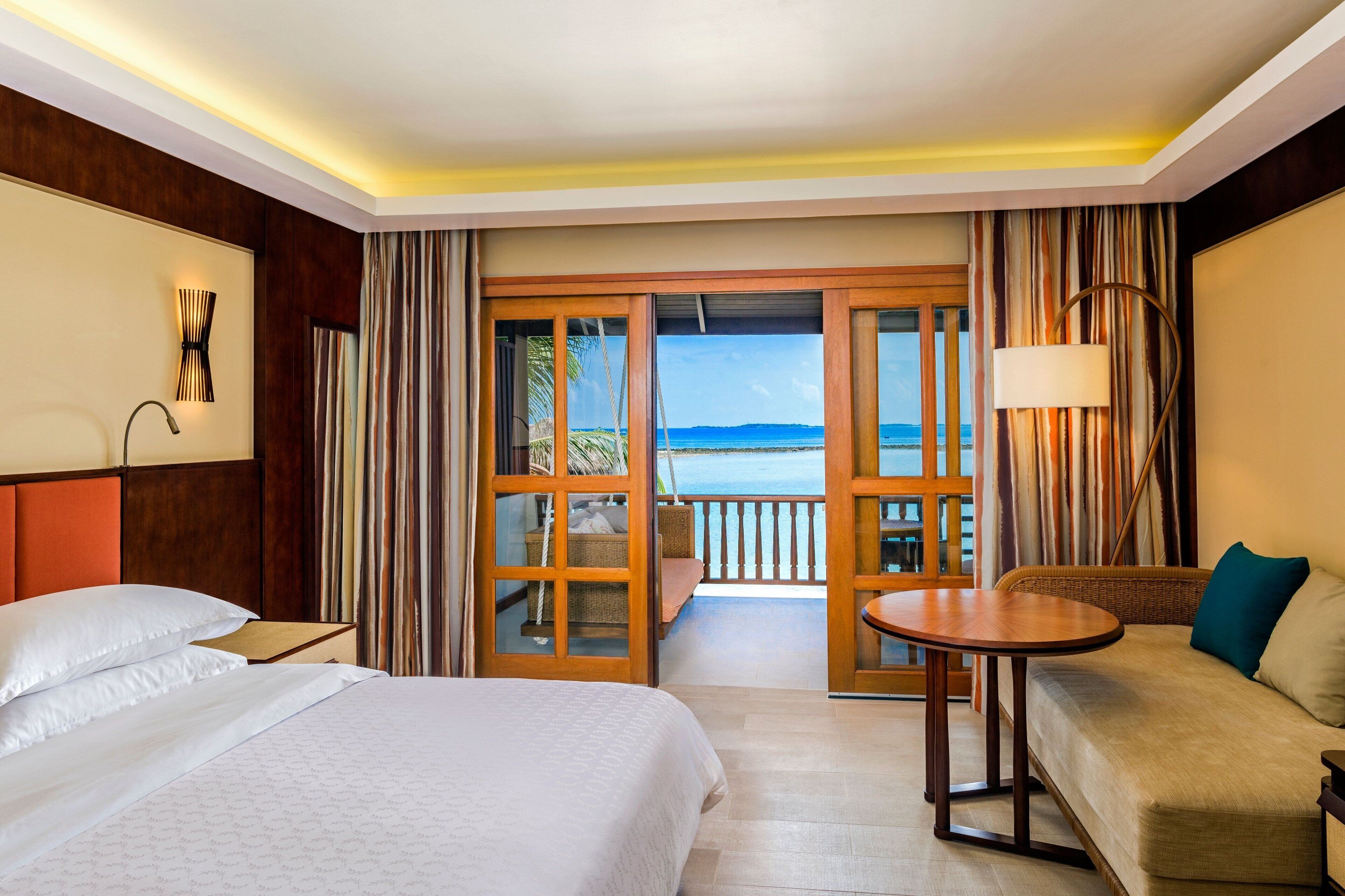 Habitación de hotel con cama, sofá, mesa y vista al océano a través de una puerta corrediza.