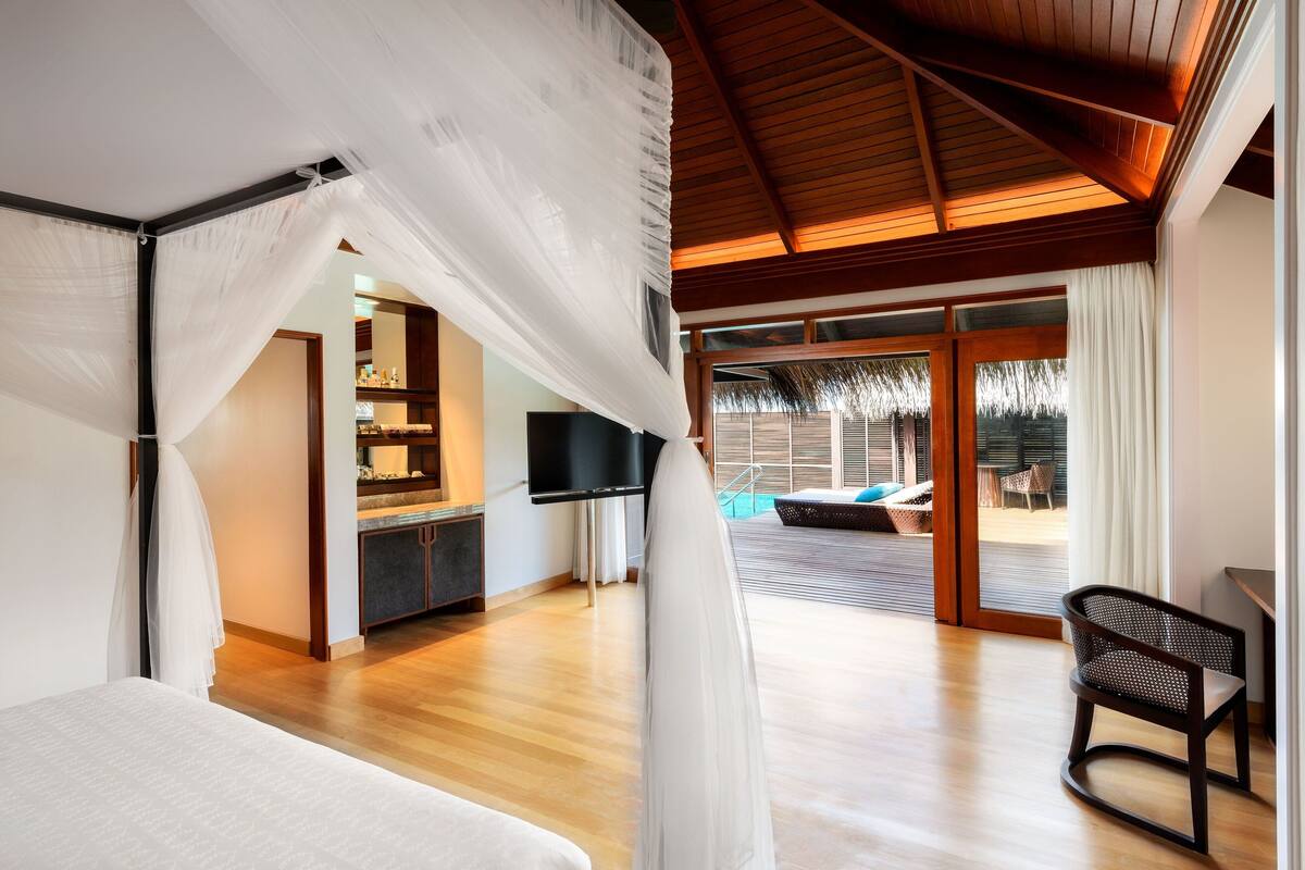 A bedroom with a canopy bed, a TV, a kitchenette, and a view of the outdoor pool area.