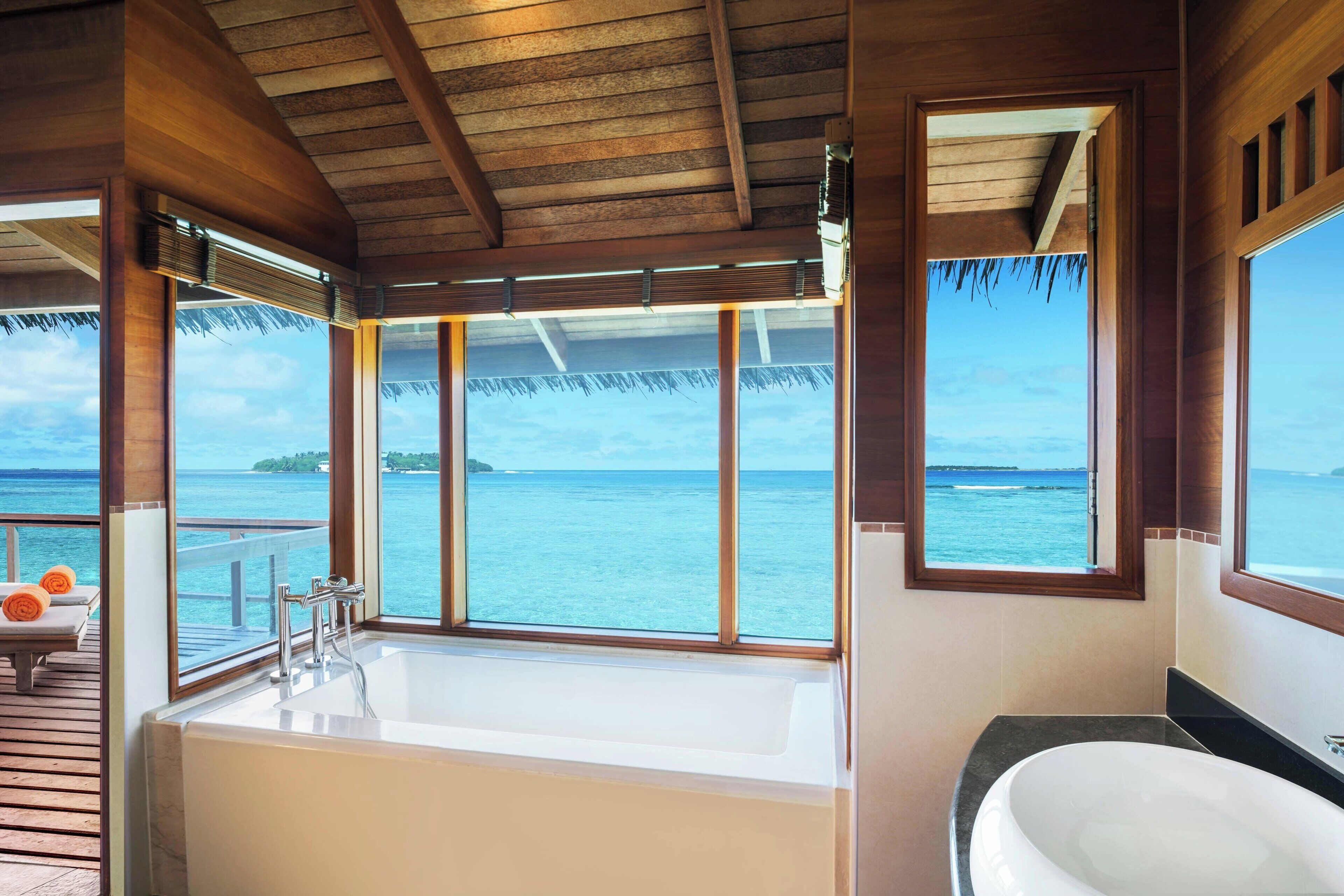 A bathroom with a bathtub and a sink, large windows with a view of the ocean, and a wooden ceiling.
