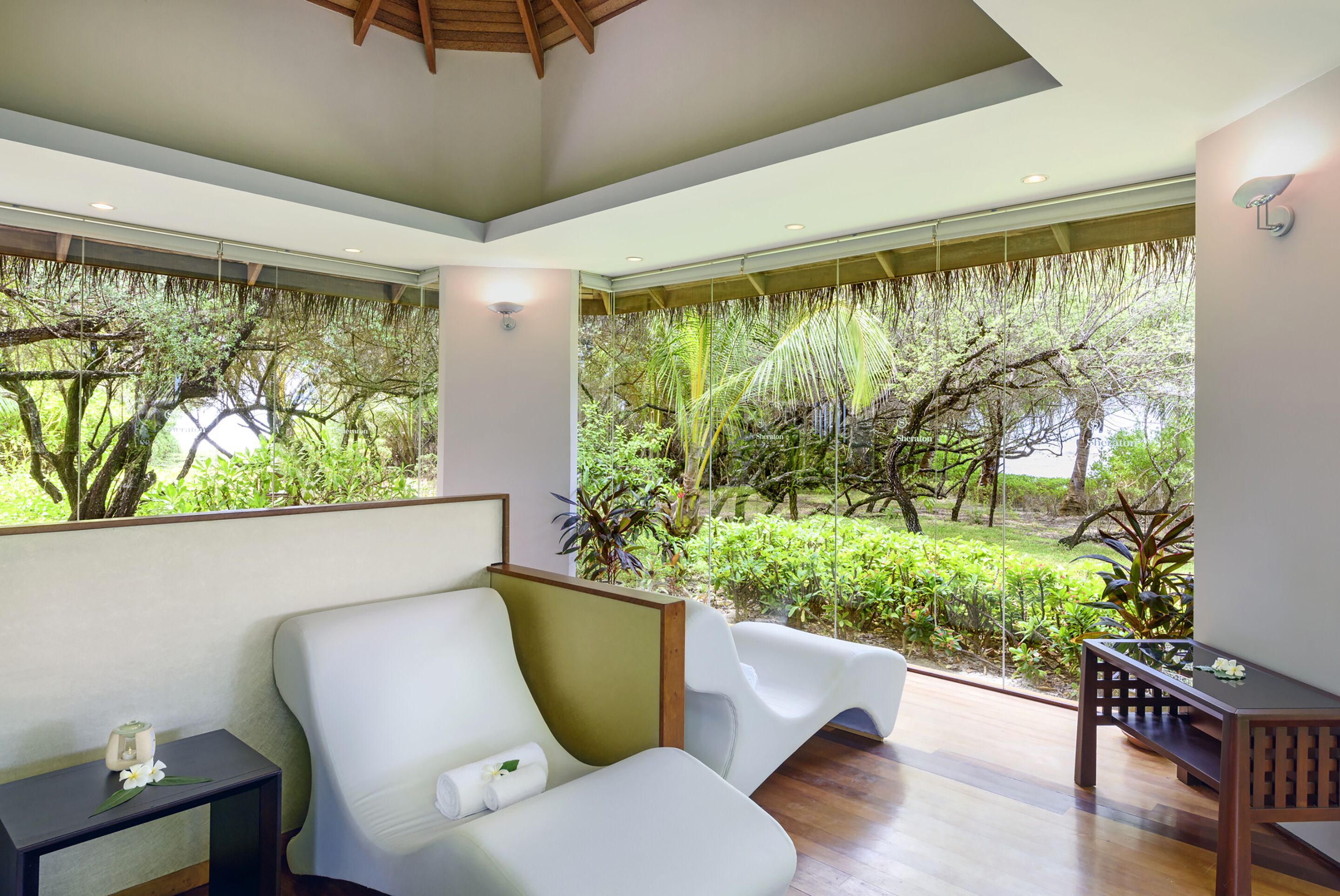 A modern room with a white sofa, a small table, and a view of a lush garden through large windows.