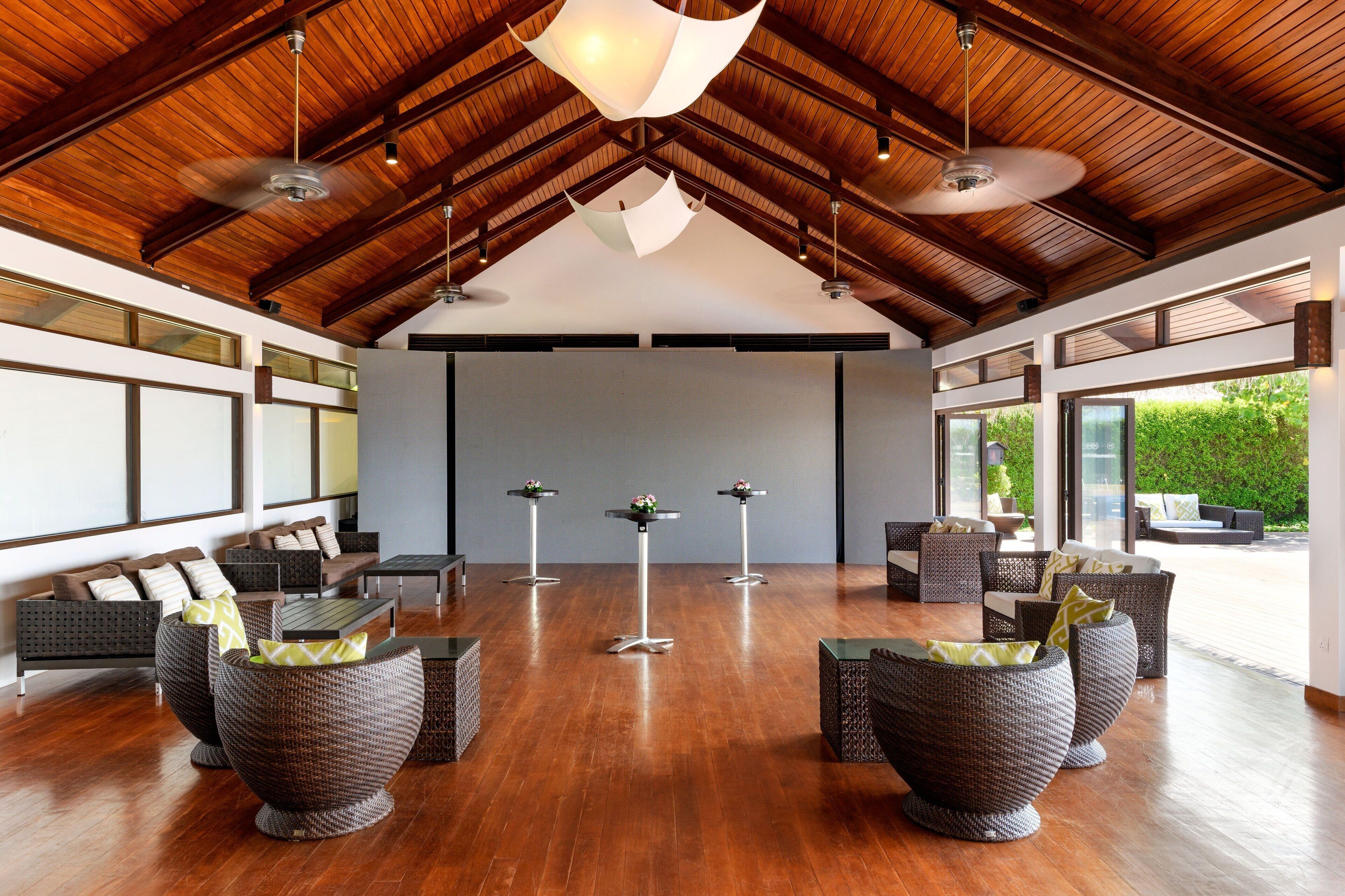 A spacious indoor area with high ceilings, wooden beams, and large windows. There are several seating areas with wicker furniture and tables, some with flower arrangements.