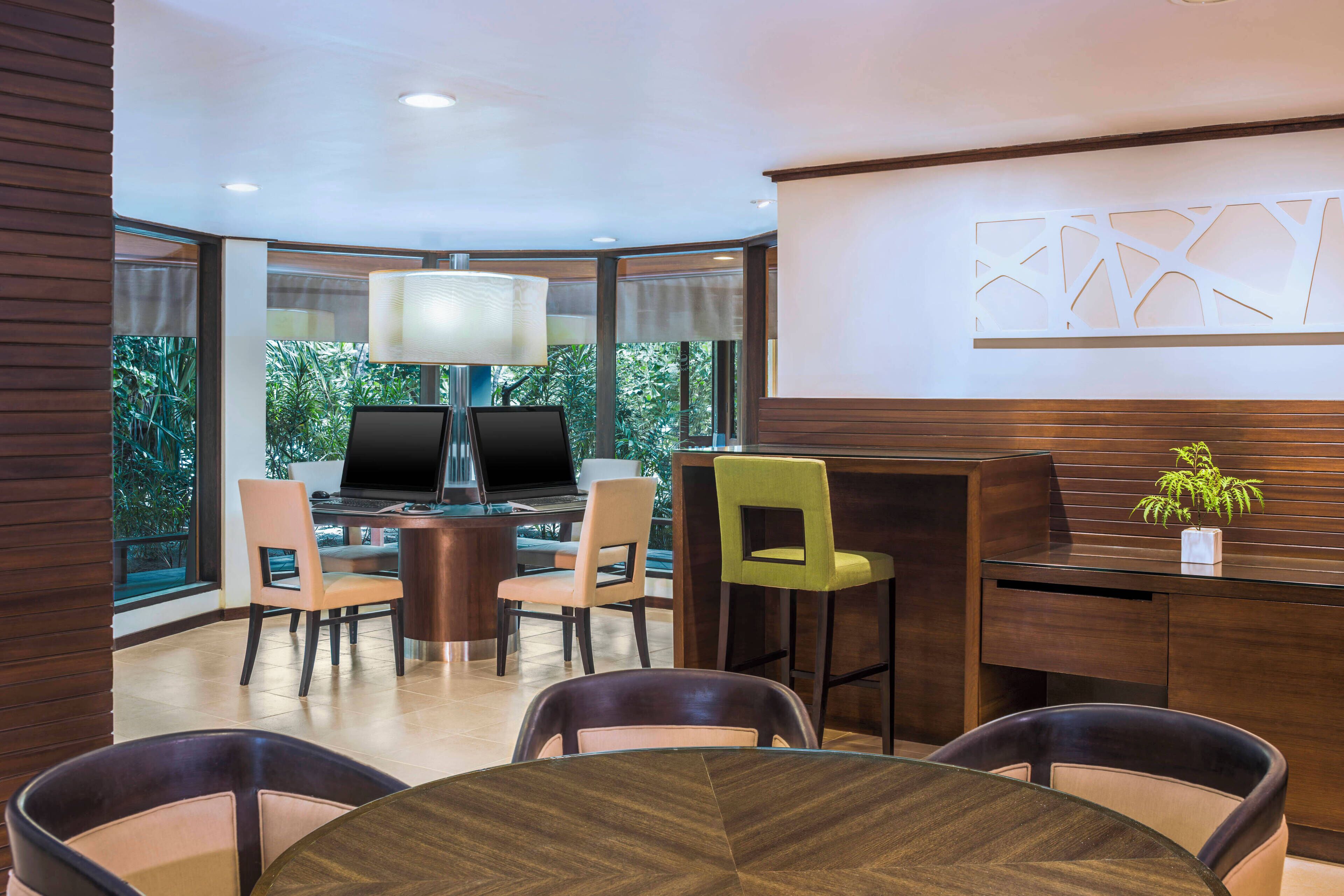 A modern hotel lobby with a round table, several chairs, a bar area, and a large window offering a view of greenery.