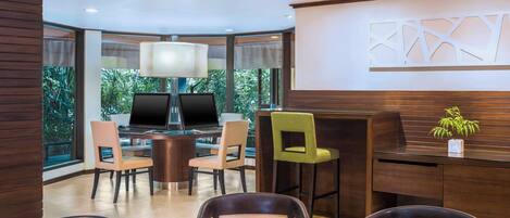 A modern hotel lobby with a round table, several chairs, a bar area, and a large window offering a view of greenery.