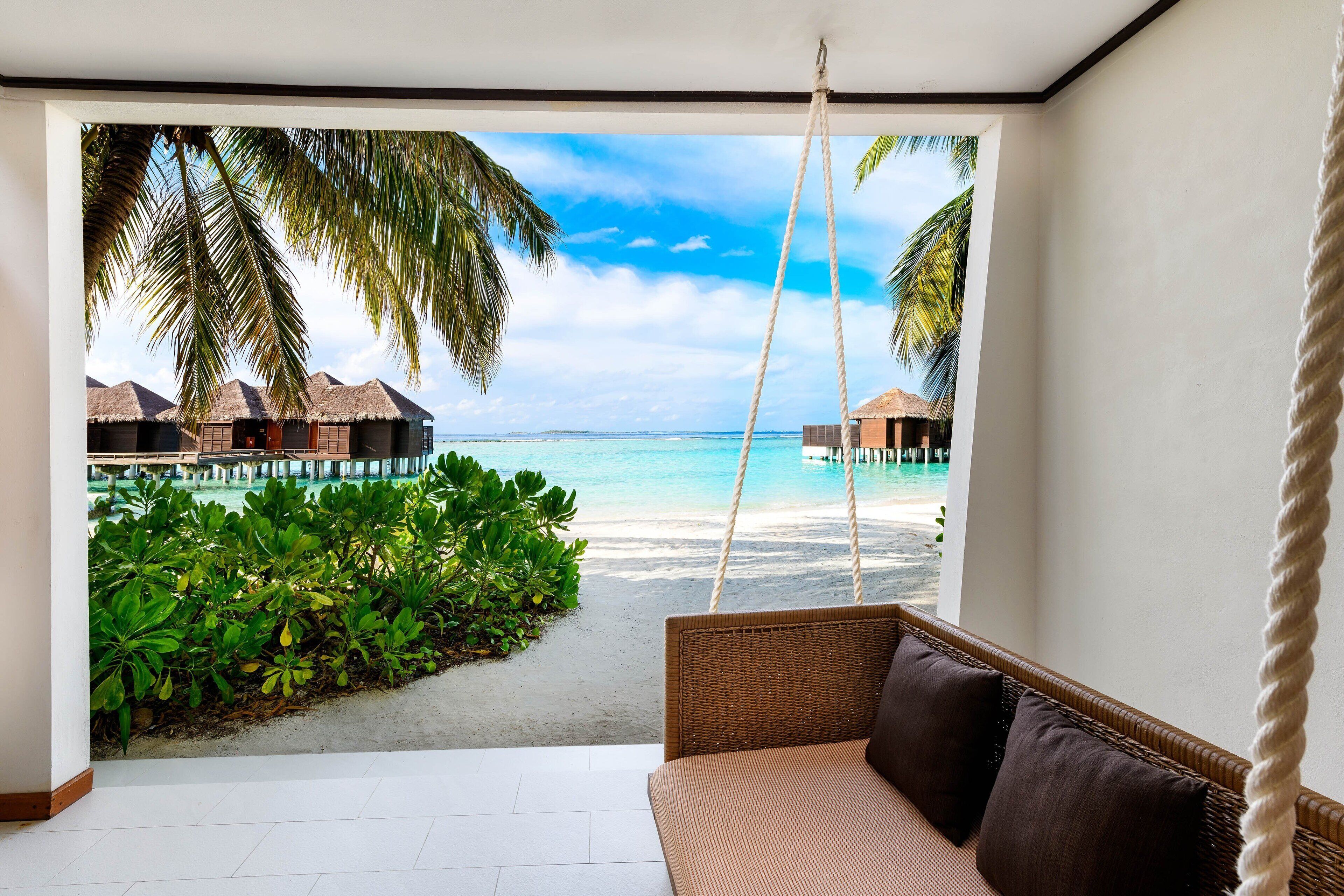 A room with a swing chair, a view of a beach, and overwater bungalows.