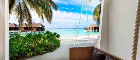 Ein Zimmer mit einer Schaukelstuhlsitzgelegenheit, Blick auf einen Strand und Bungalows über dem Wasser.
