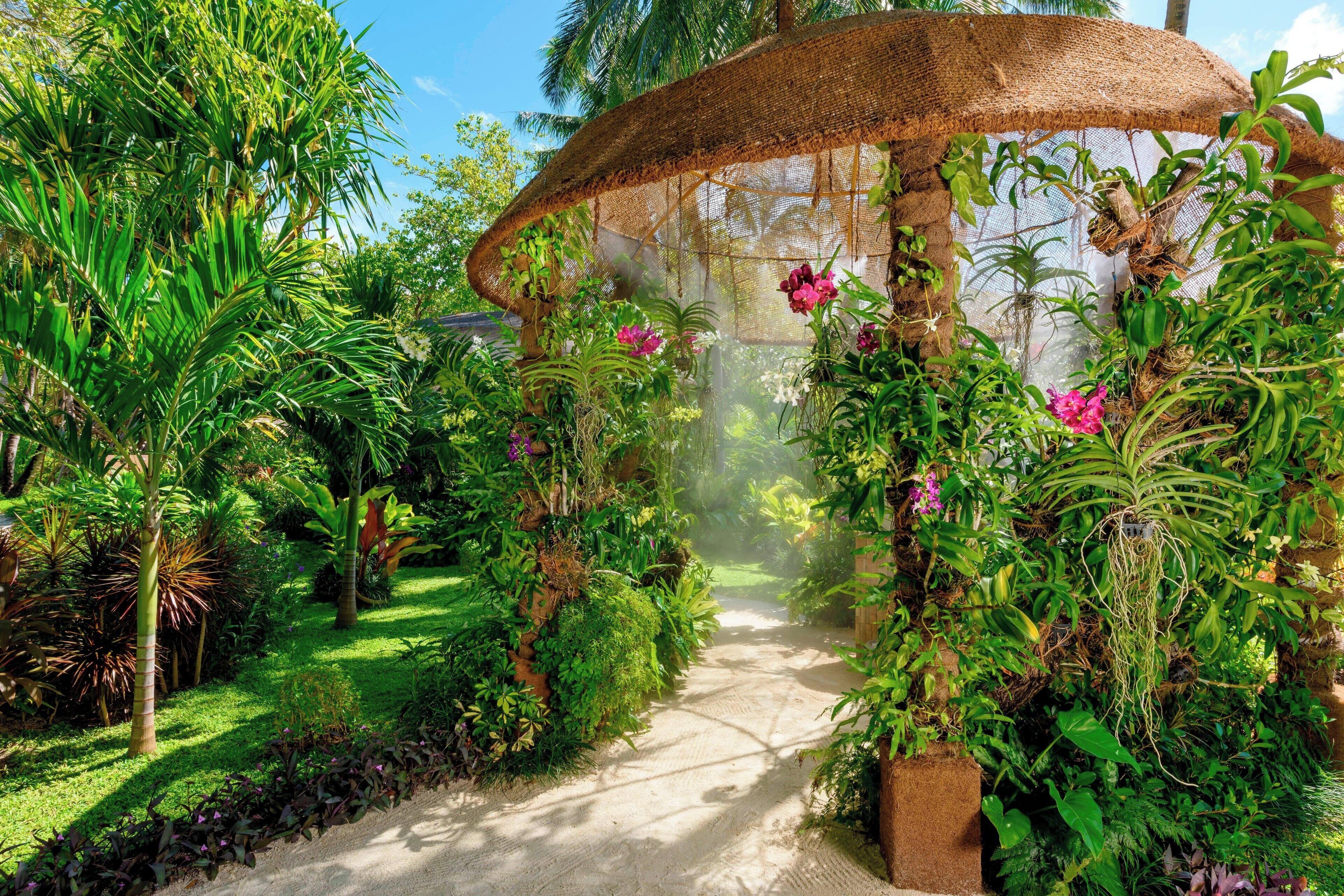 Un sendero en un jardín con una estructura de paja, rodeado de exuberantes plantas y flores tropicales.