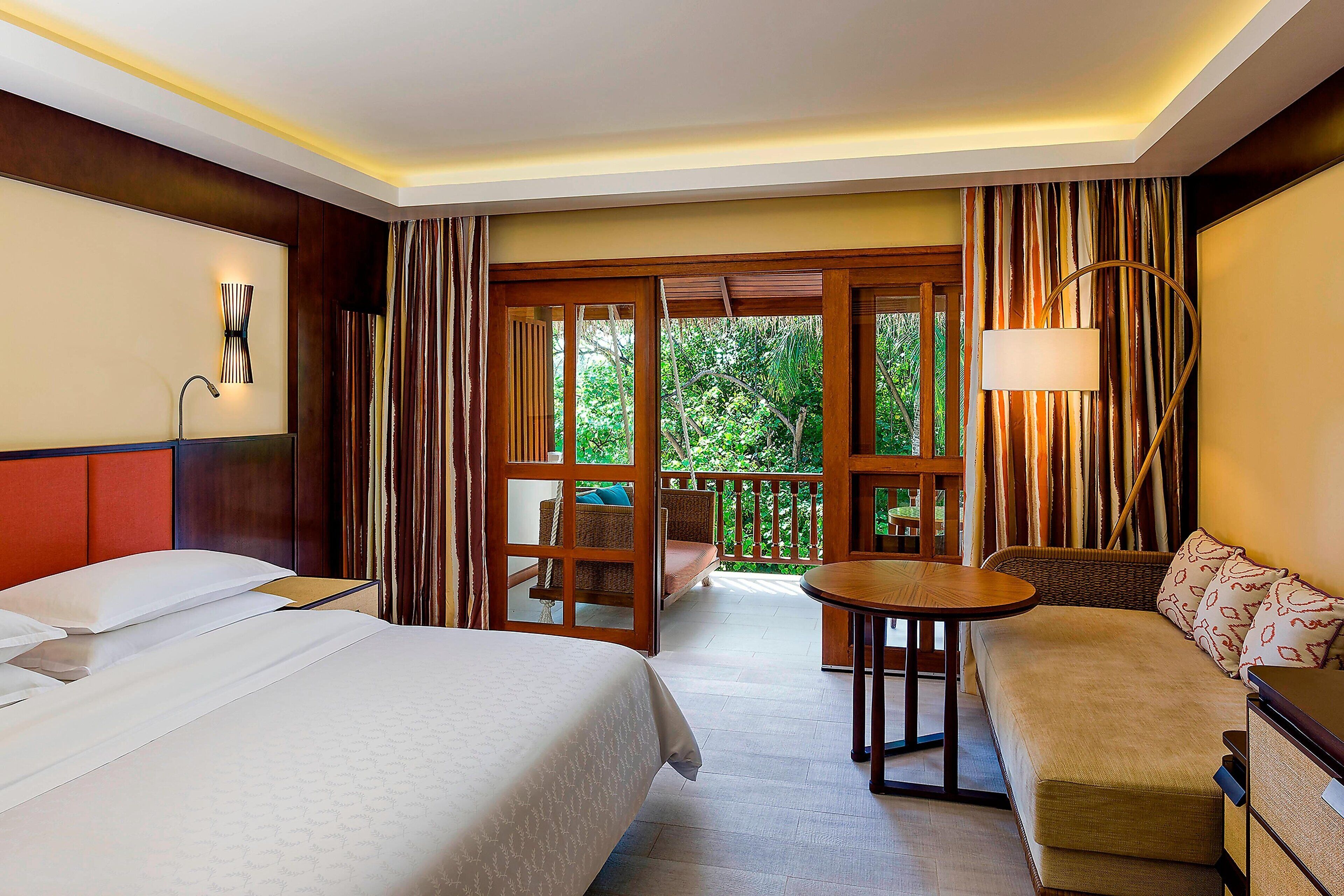 A hotel room with two beds, a sofa, a small table, and a balcony with a view of greenery.