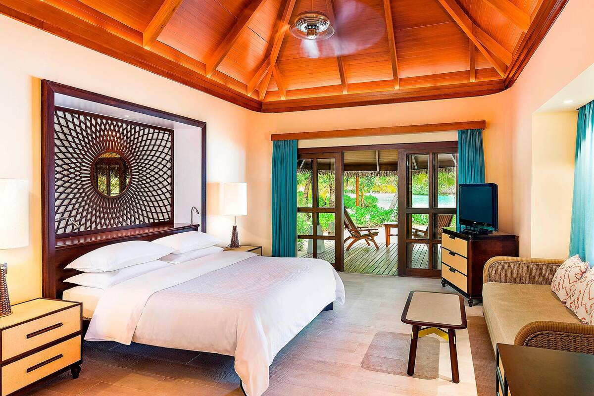 A hotel room with a large bed, a TV, a sofa, and a balcony with a view of greenery.