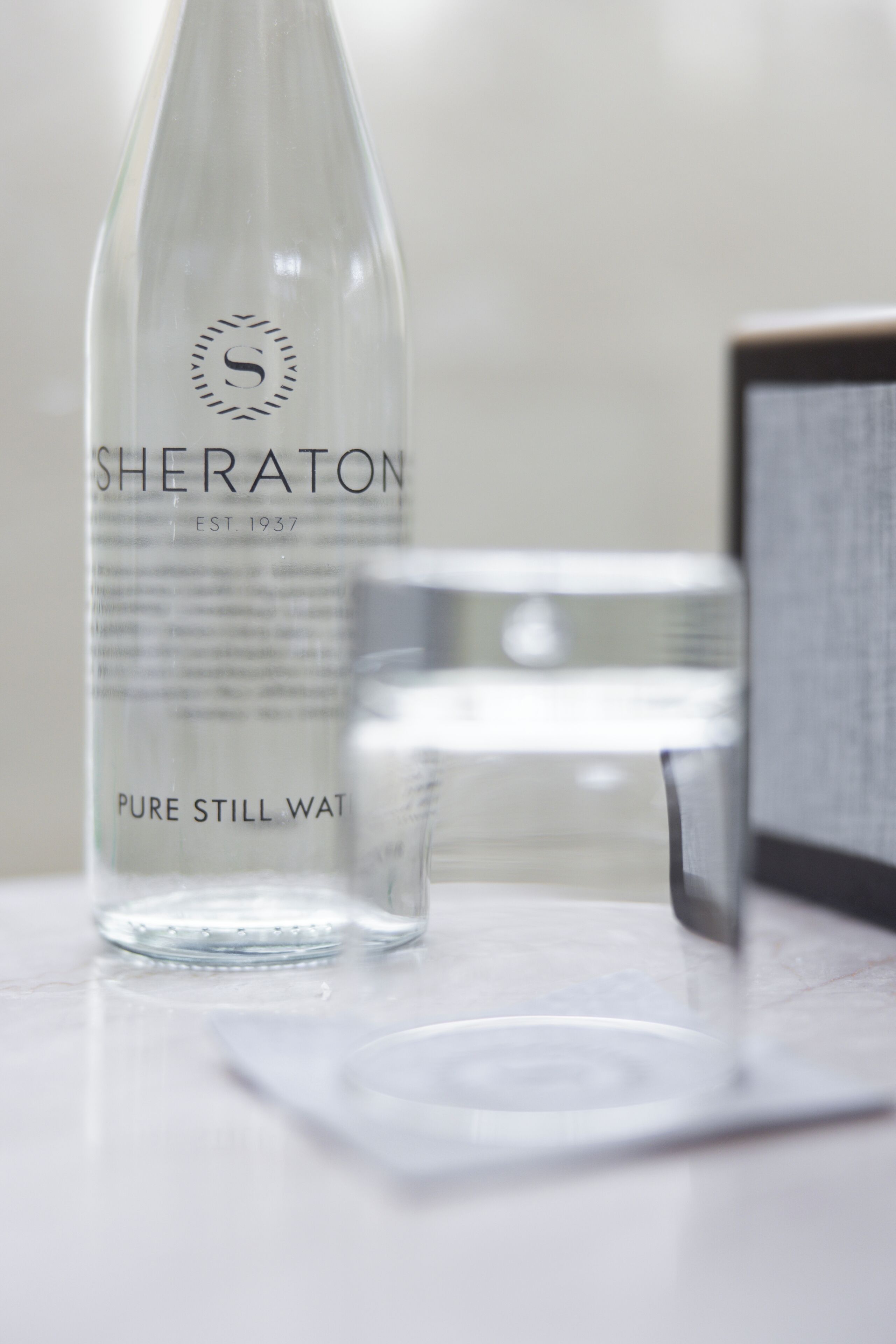 A clear glass bottle labeled 'SHERATON EST. 1937 PURE STILL WATER' on a marble surface.