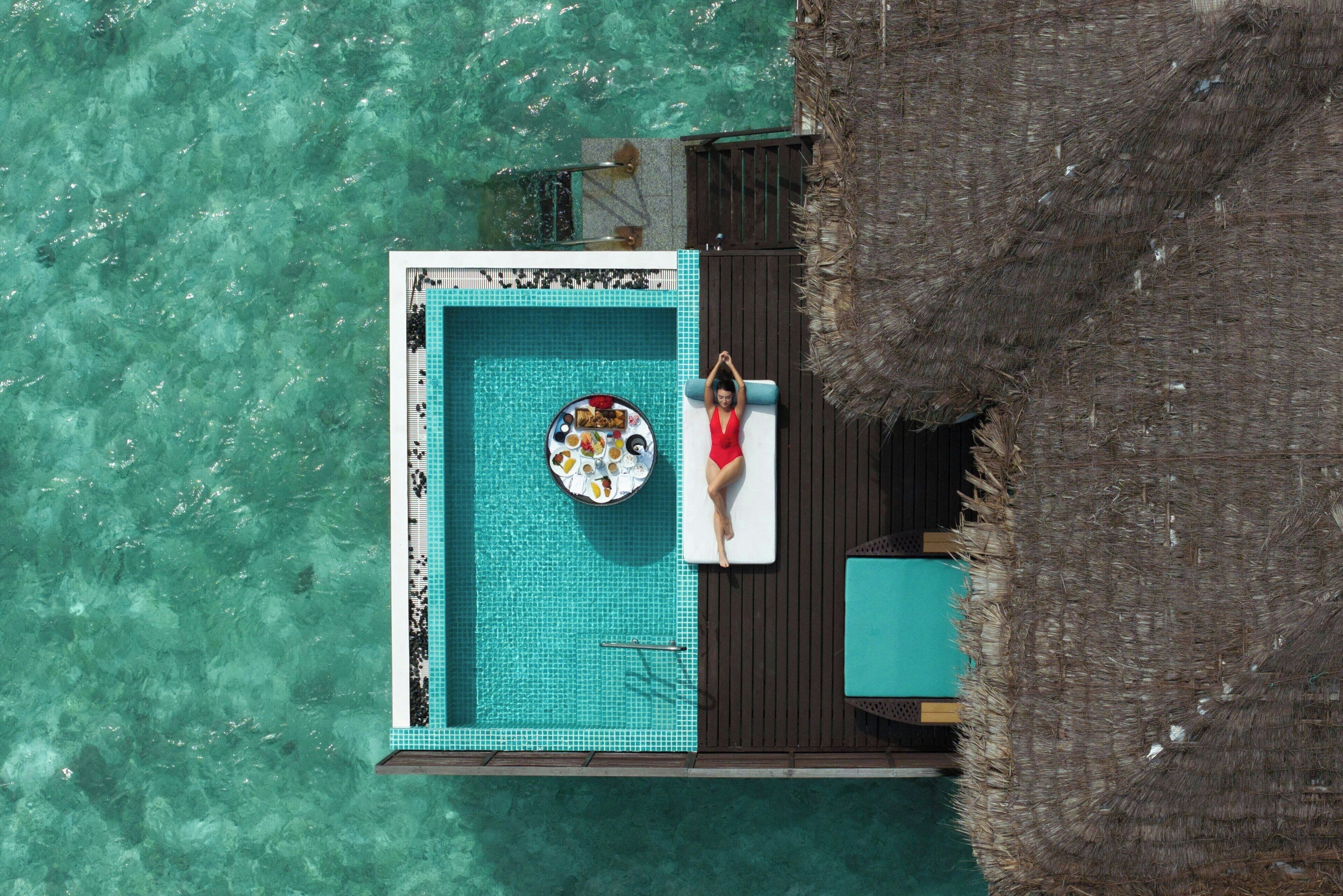 An aerial view of a private pool area with a person relaxing on a lounge chair, surrounded by a wooden deck and a thatched roof structure.