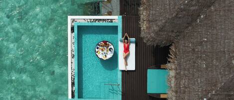 An aerial view of a private pool area with a person relaxing on a lounge chair, surrounded by a wooden deck and a thatched roof structure.