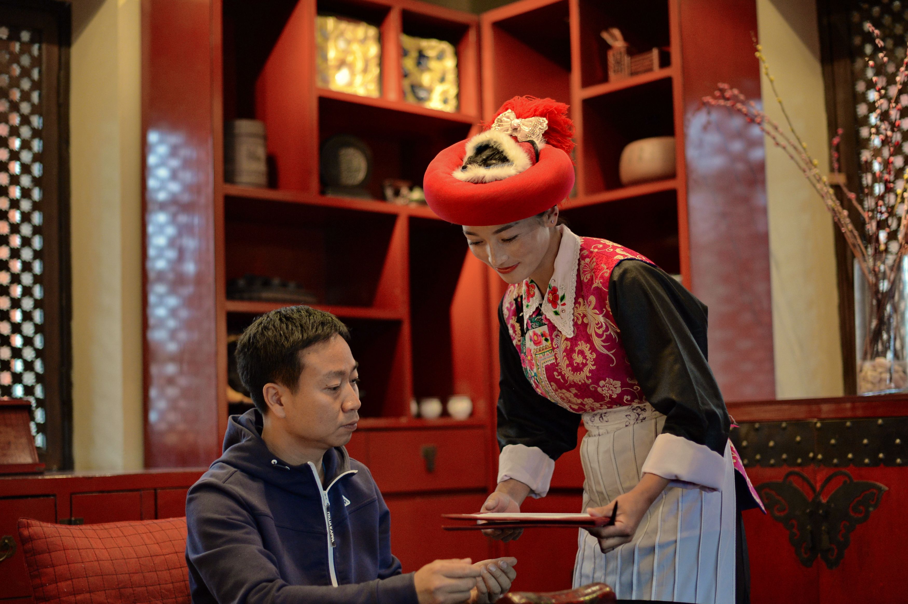 A person in traditional attire standing next to a seated individual in a room with red decor and shelves containing various items.