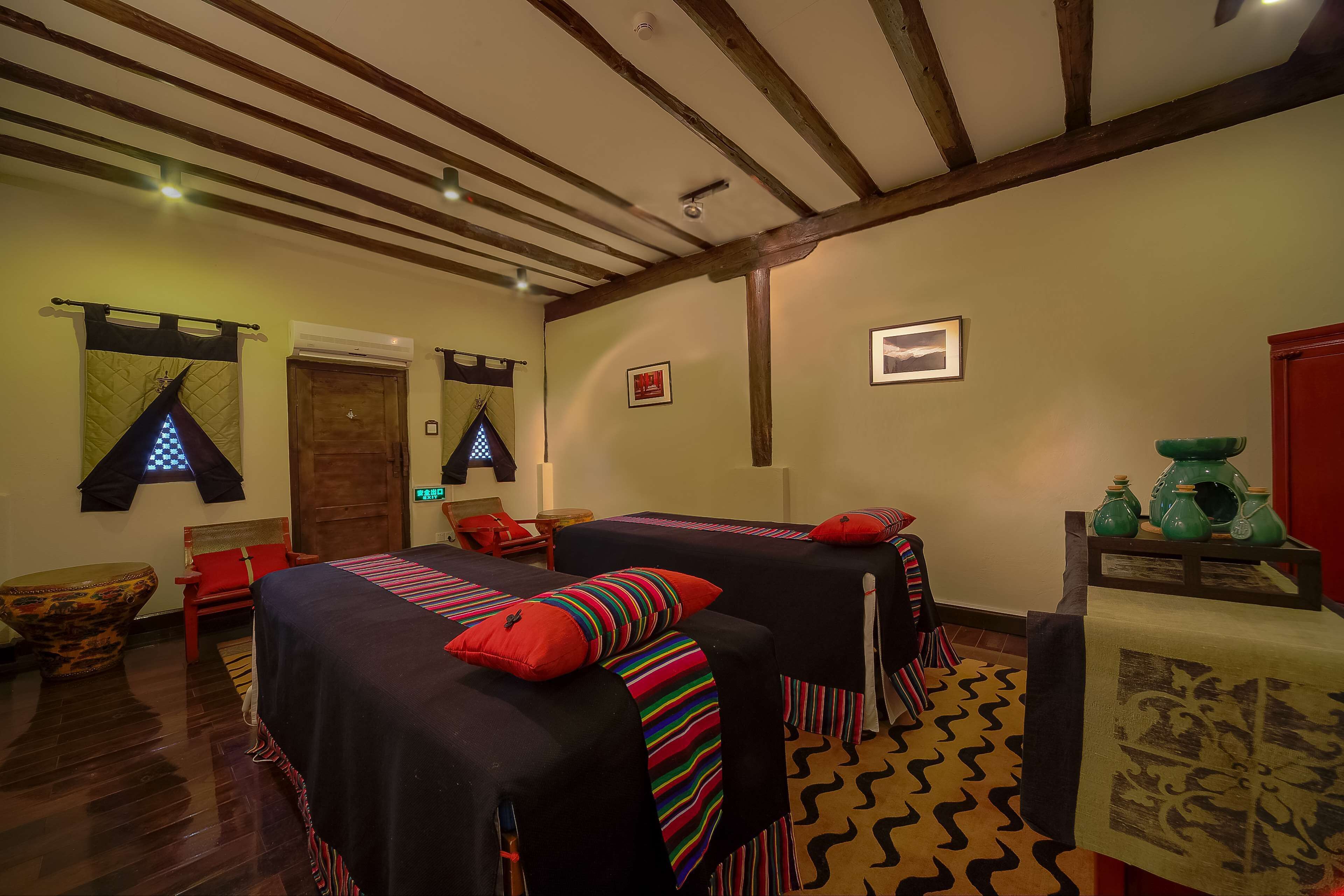 A room with two beds, a wooden ceiling, a small table with green vases, and a patterned rug.