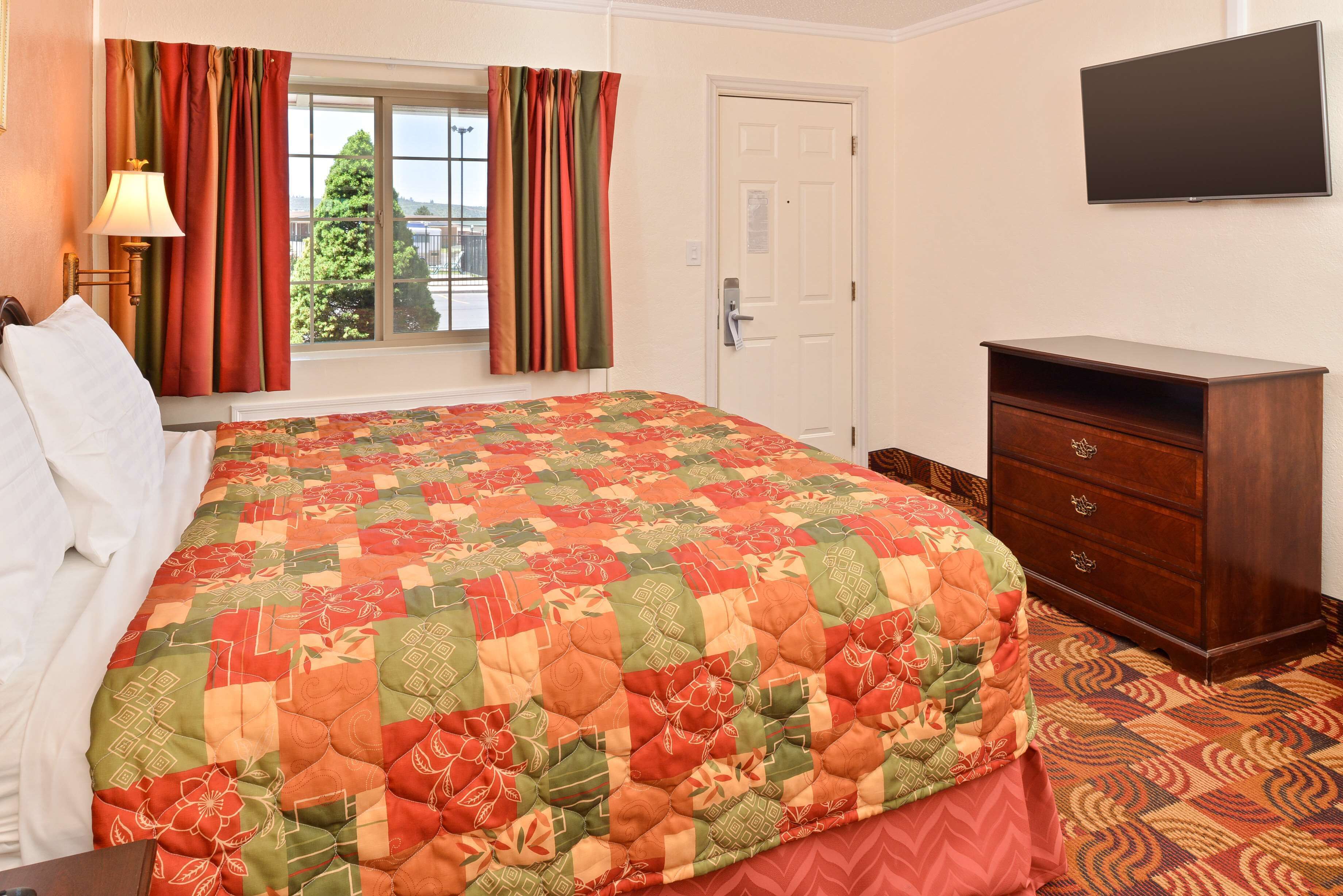 A hotel room with a large bed, a dresser, a television on the wall, and a window with curtains.