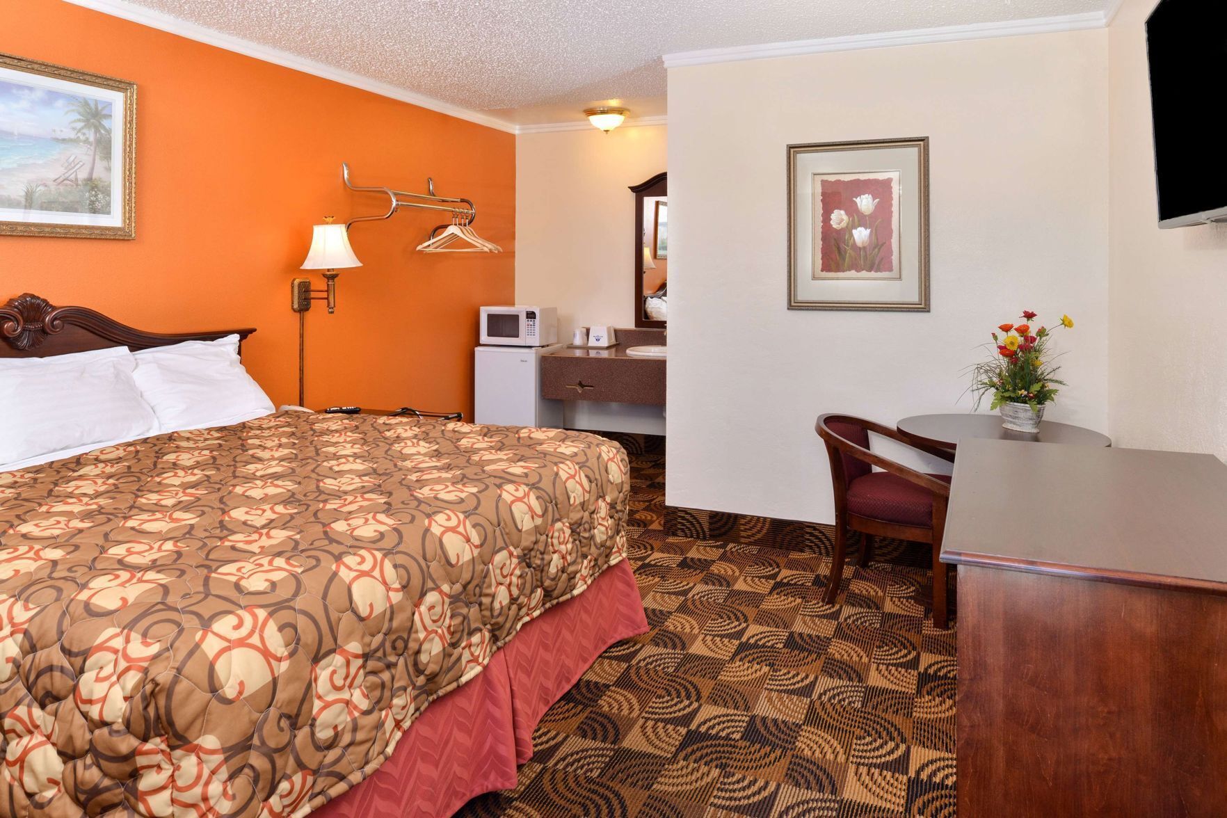 Habitación de hotel con cama, escritorio, silla, microondas, lámpara y un cuadro en la pared.
