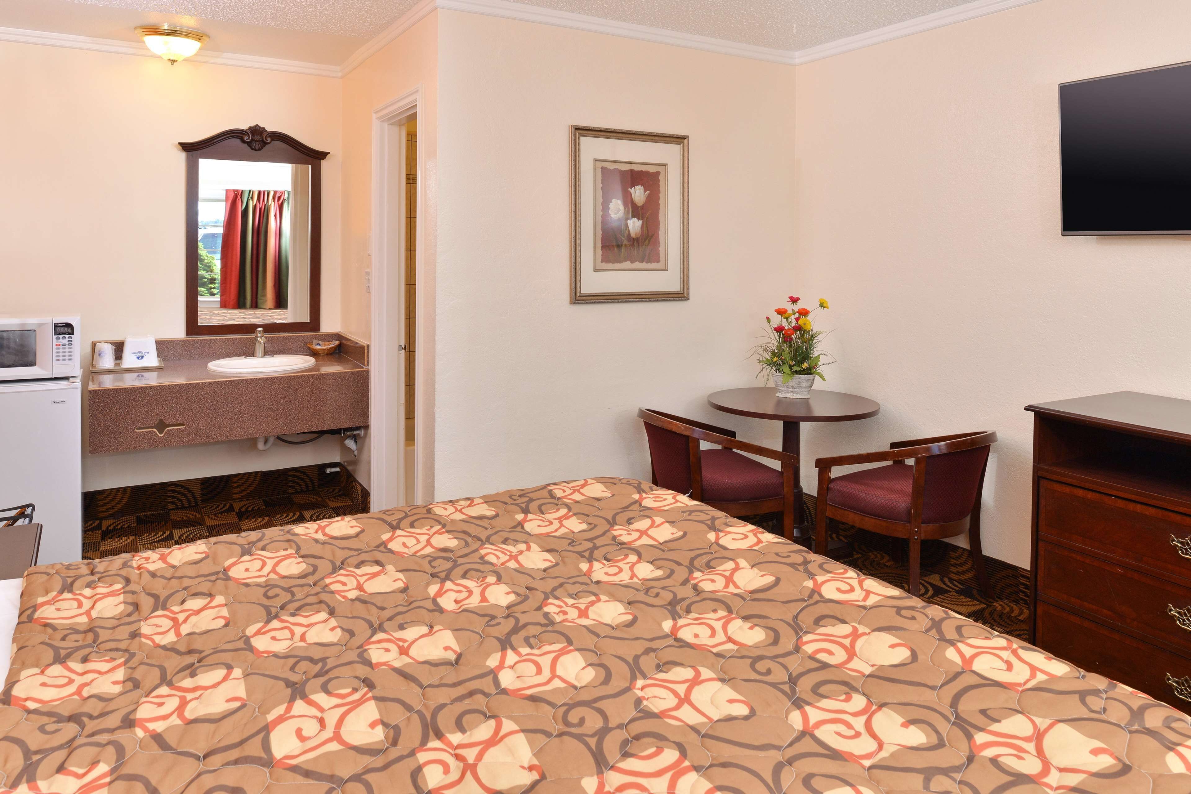 A hotel room with a bed, a bathroom with a sink and mirror, a TV, a small table with chairs, and a dresser.