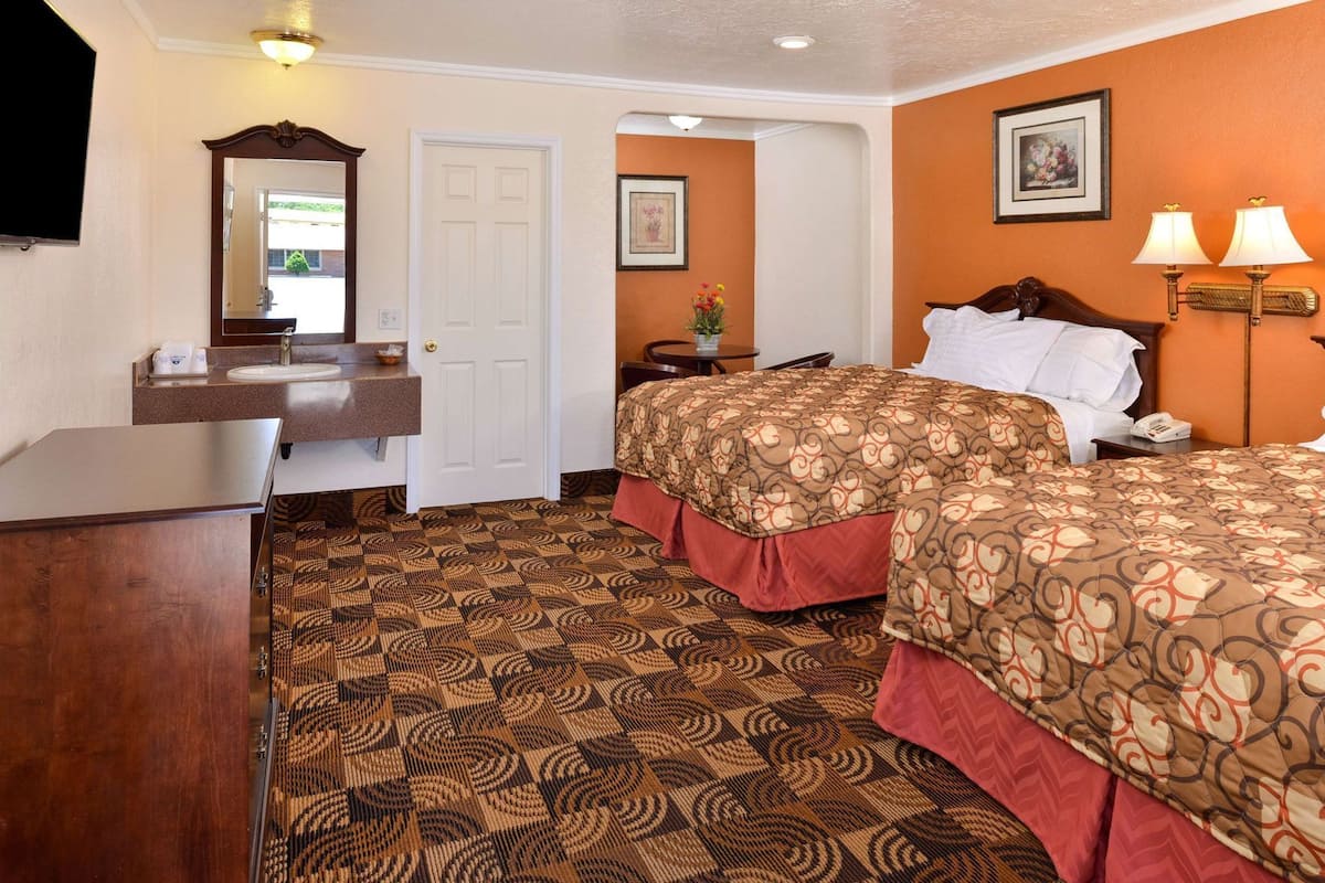 A hotel room with a bed, a desk, a mirror, a door, and a wall with a framed picture.