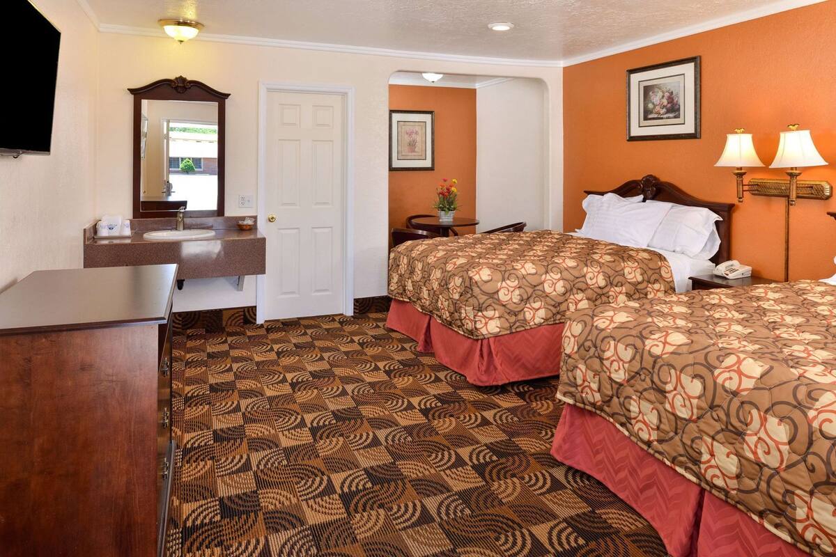 A hotel room with a bed, a desk, a mirror, a door, and a wall with a framed picture.