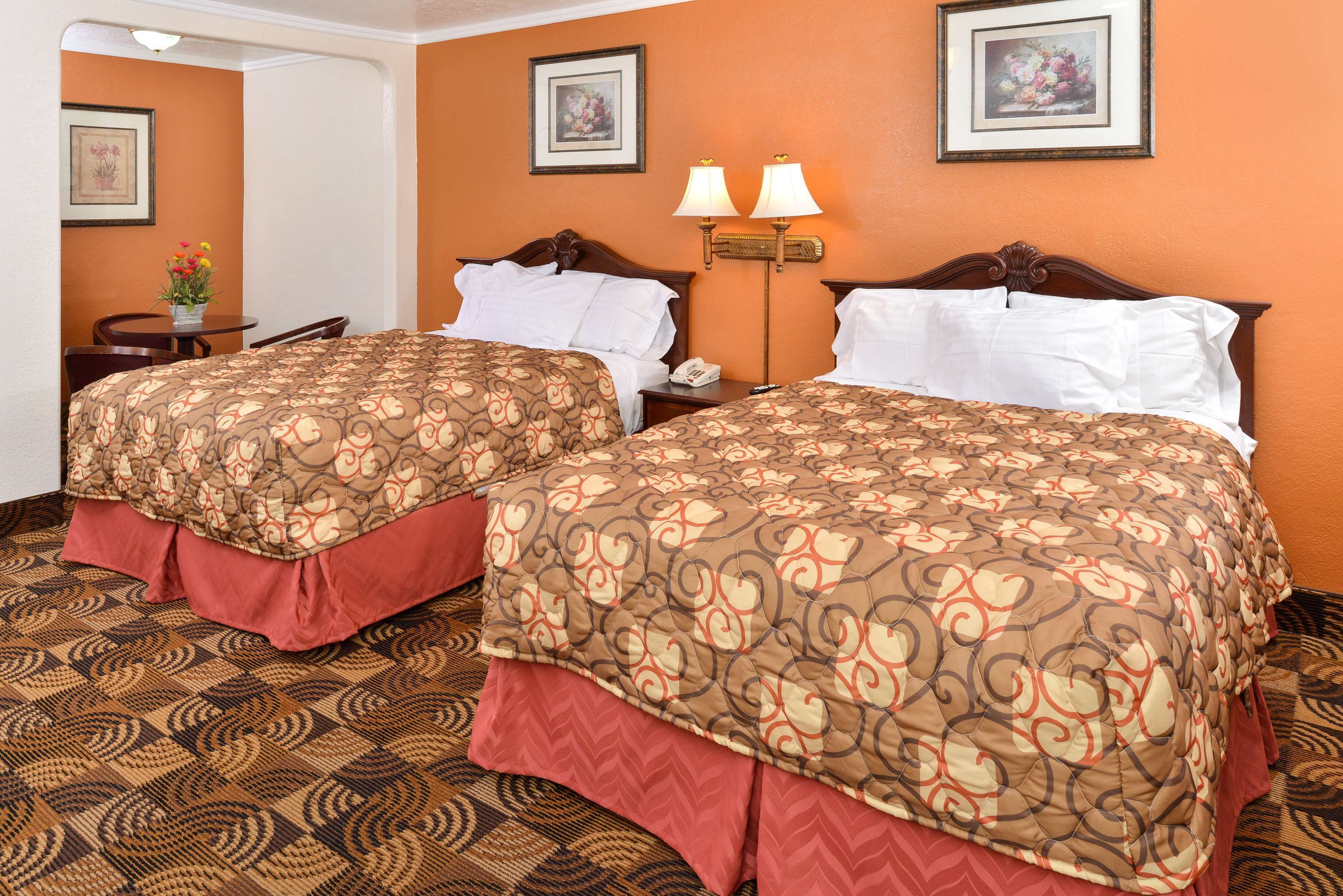 A hotel room with two beds, a desk, and a chair. There are two lamps on the wall and a framed picture above the desk.