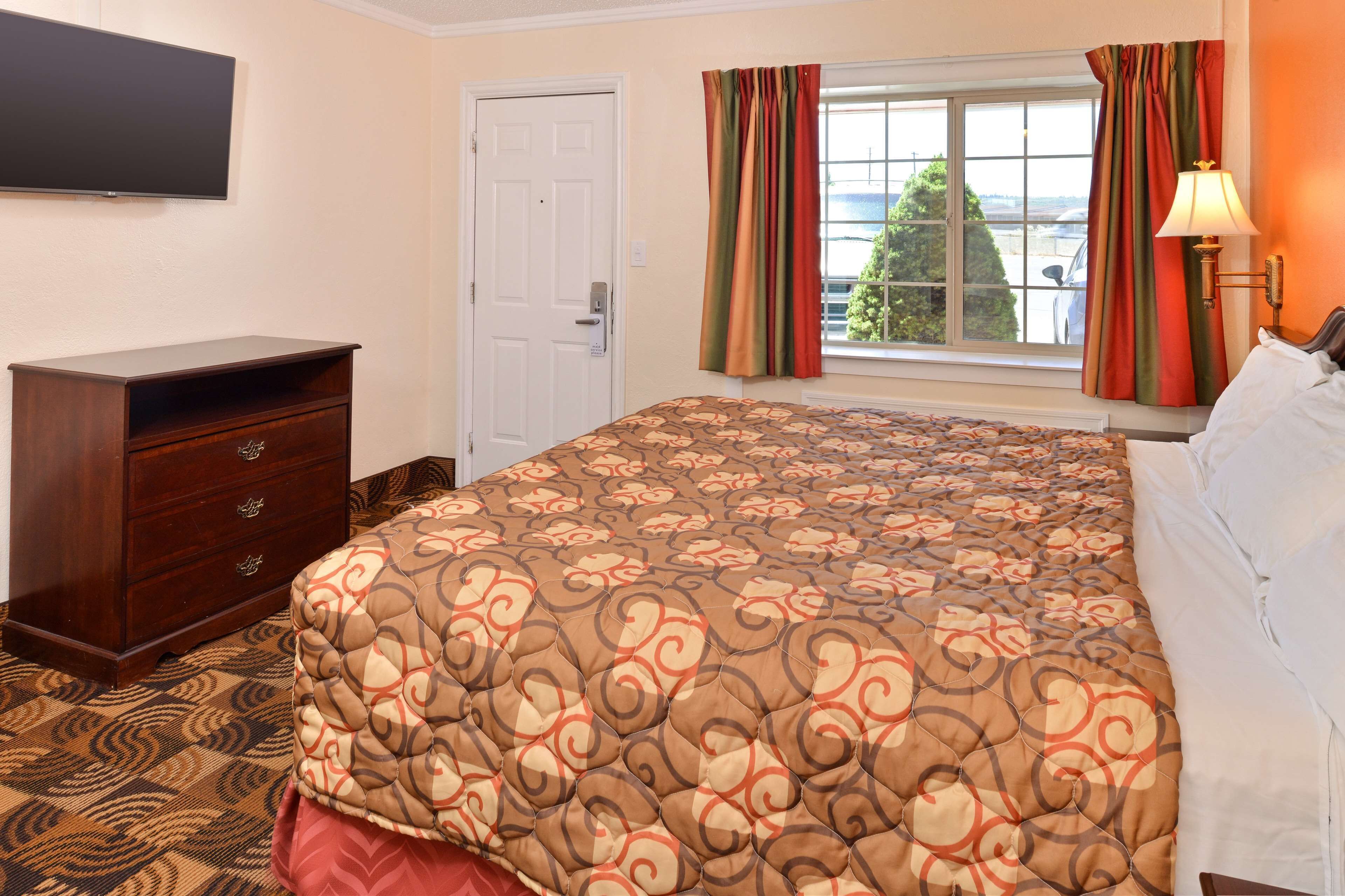 A hotel room with a bed, a dresser, a television on the wall, a window with curtains, and a lamp on the nightstand.