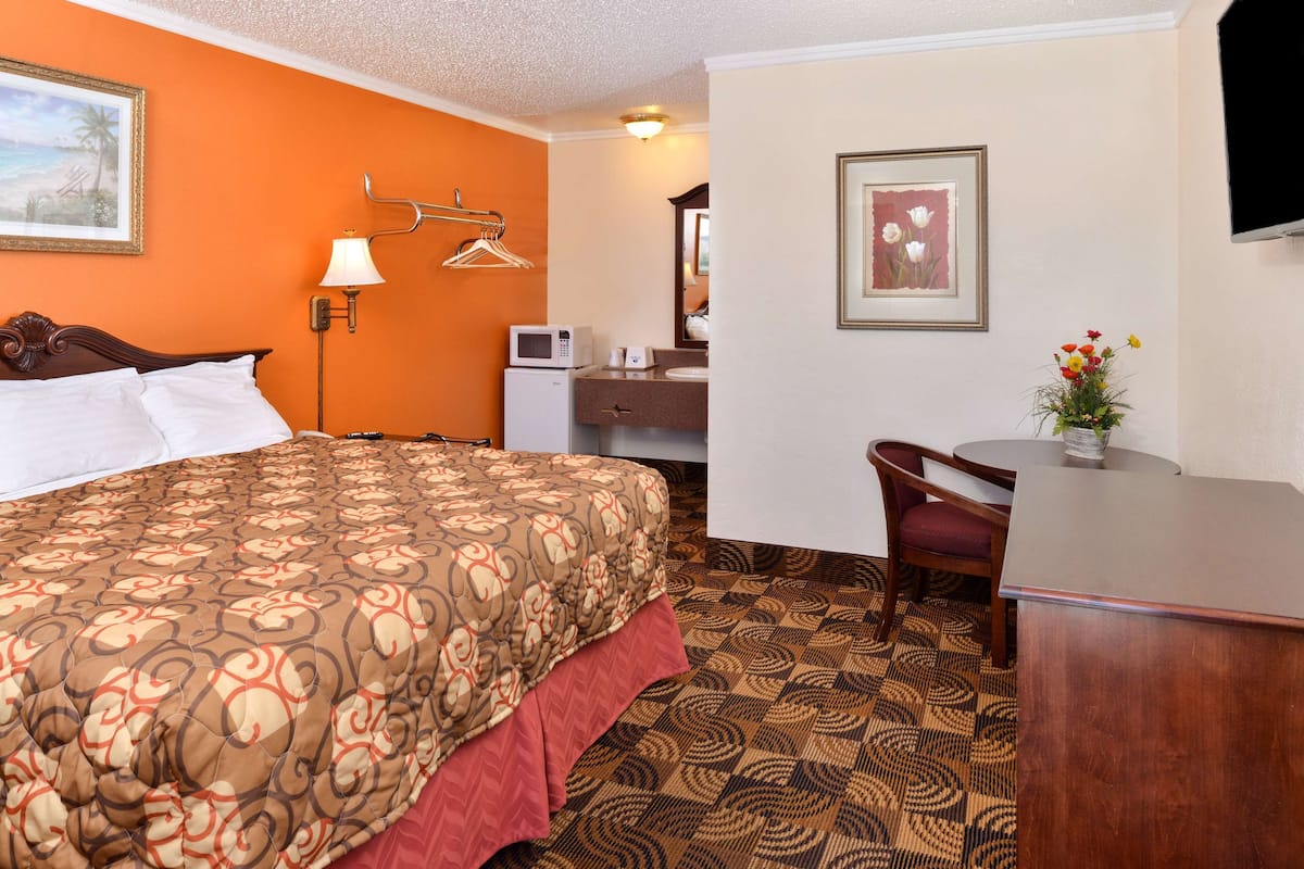 A hotel room with a bed, a desk, a chair, a microwave, and a small table with a vase of flowers.