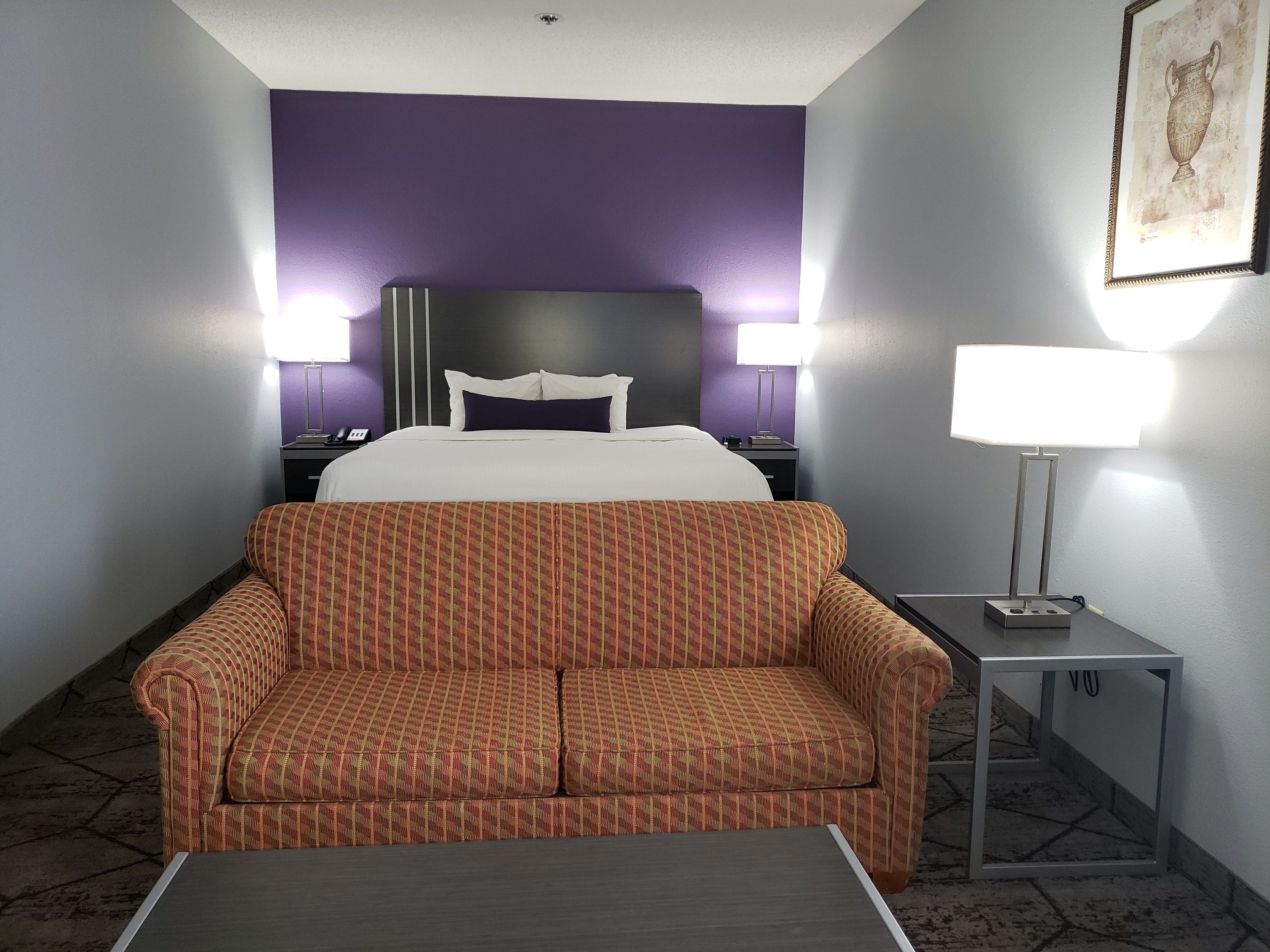 A hotel room with a bed, two bedside lamps, a sofa, a small table, and a framed picture on the wall.