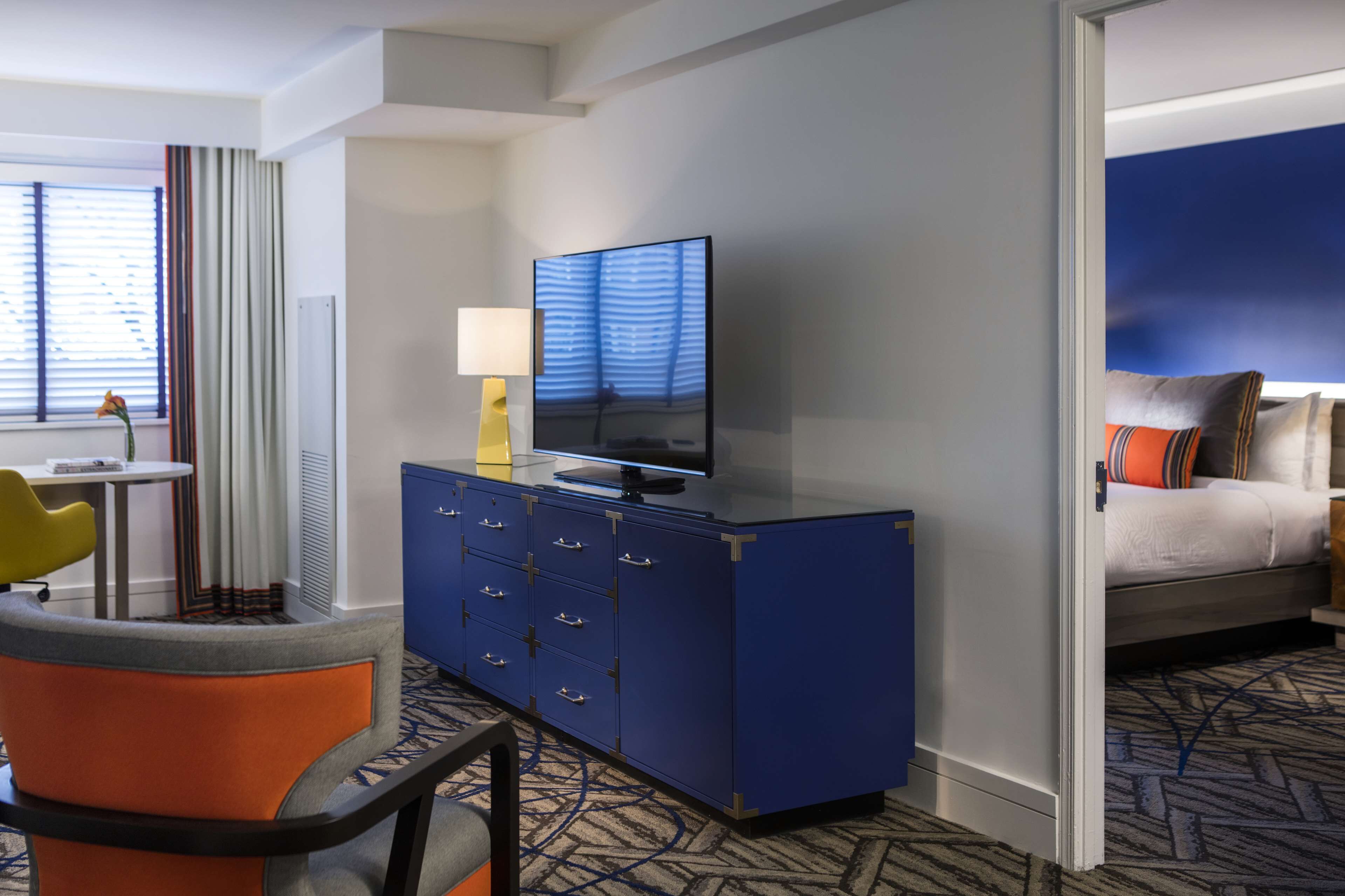 Une chambre d’hôtel avec un lit, un bureau doté d’une lampe, un téléviseur posé sur une commode bleue et une fenêtre avec des rideaux.