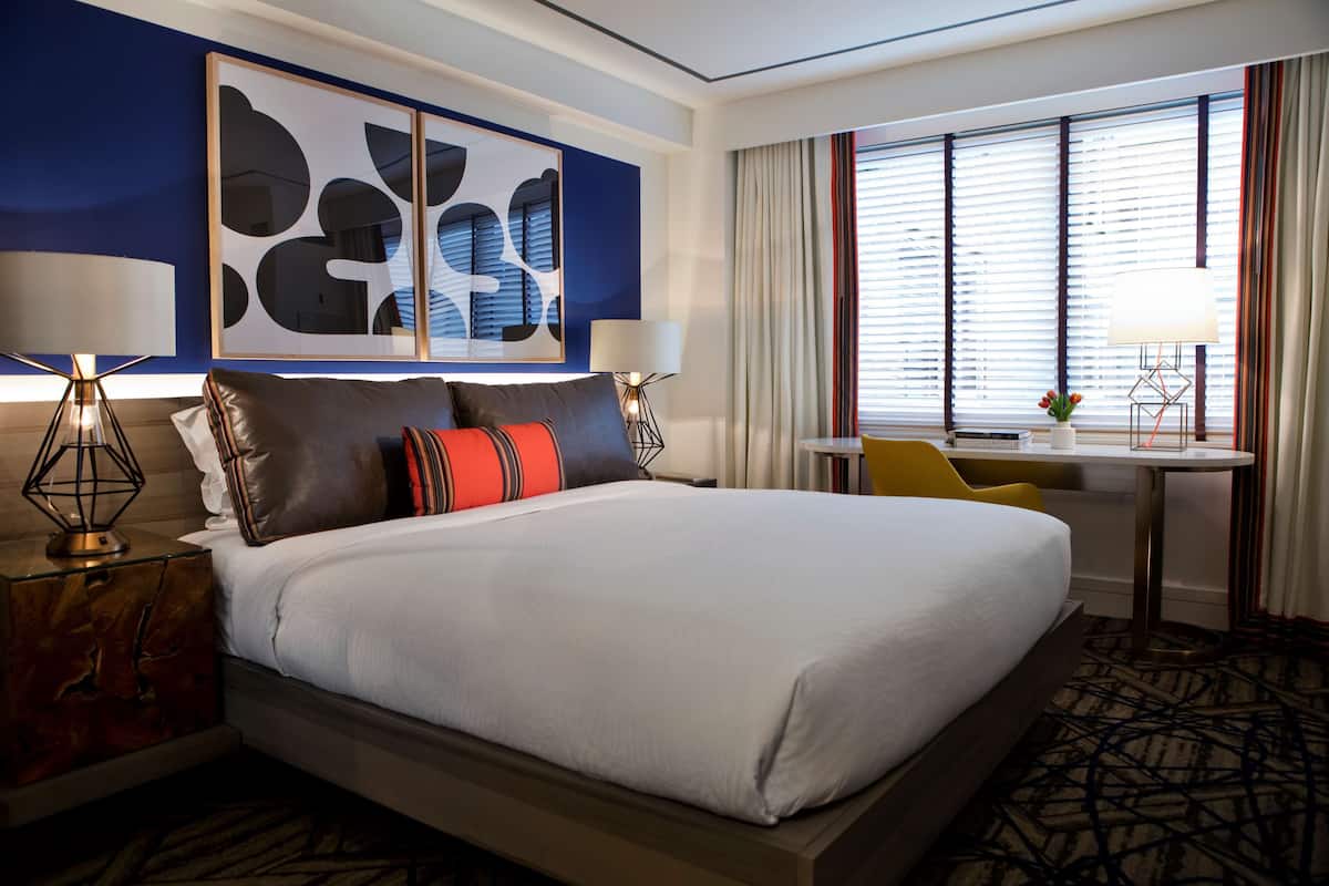A hotel room with a large bed, two bedside tables with lamps, a desk with a chair, and a window with blinds.