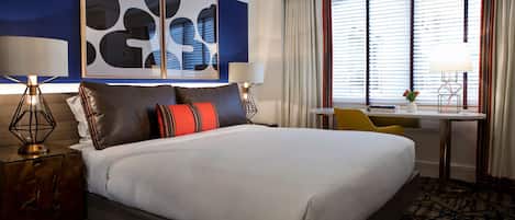 A hotel room with a large bed, two bedside tables with lamps, a desk with a chair, and a window with blinds.