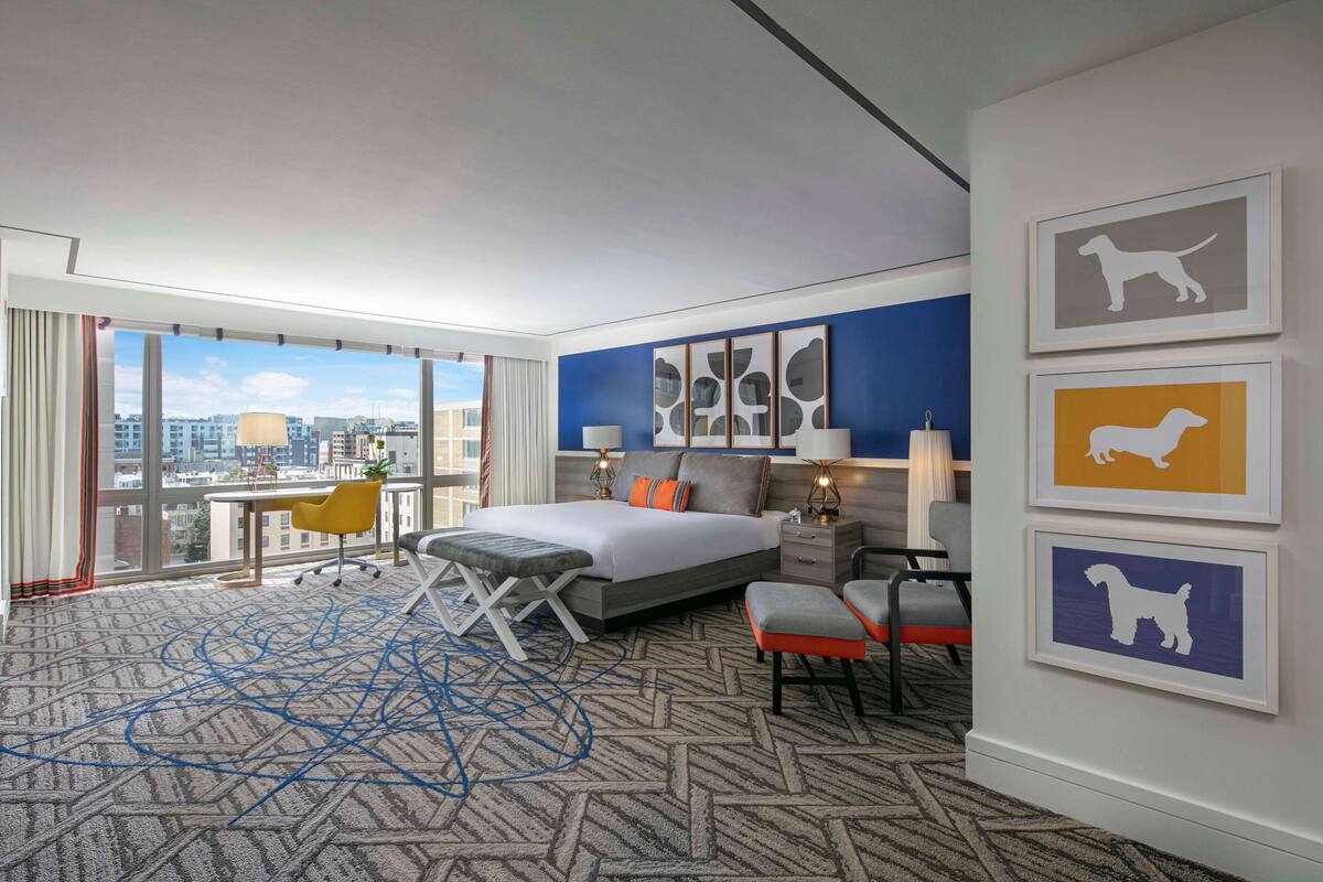 A modern hotel room with a large bed, a desk, a chair, and a view of the city.