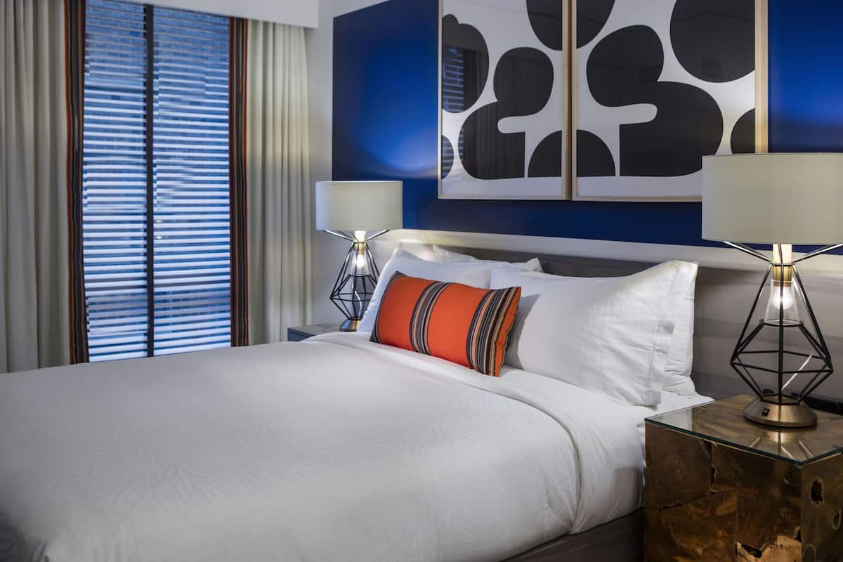A hotel room with a large bed, two bedside tables with lamps, a window with blinds, and a wall with abstract art.