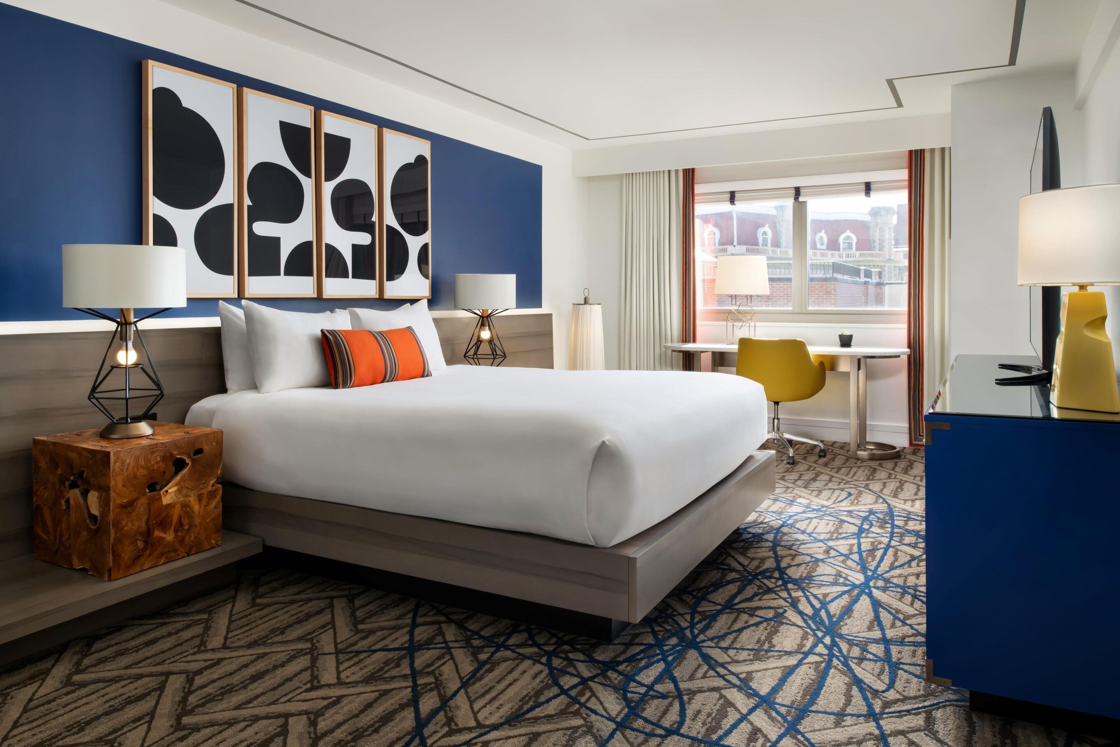 Habitación de hotel con una cama grande, lámparas de noche, una mesita de madera, un escritorio con una silla y vistas a la ciudad desde la ventana.