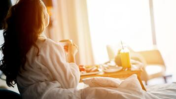 A person in a white bathrobe sitting on a bed with a breakfast tray containing a glass of orange juice, a plate of food, and a cup.