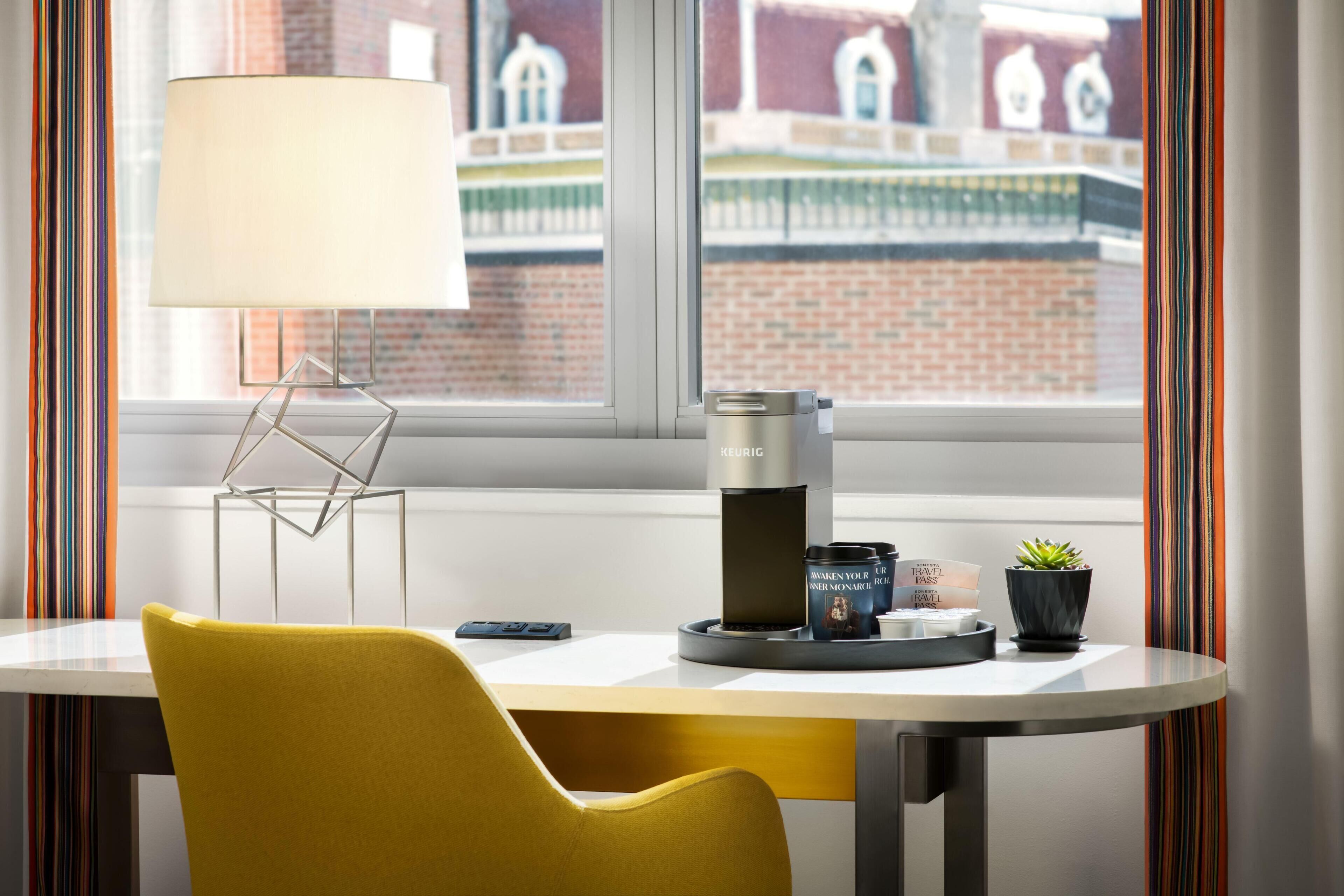 A modern room with a yellow chair, a table with a coffee maker, a lamp, and a view of a brick building.