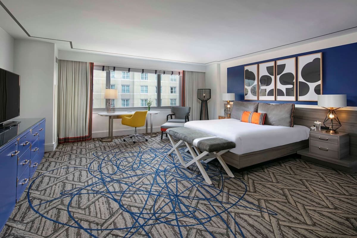 A hotel room with a bed, bedside tables, a desk, a chair, a TV, and a large window.
