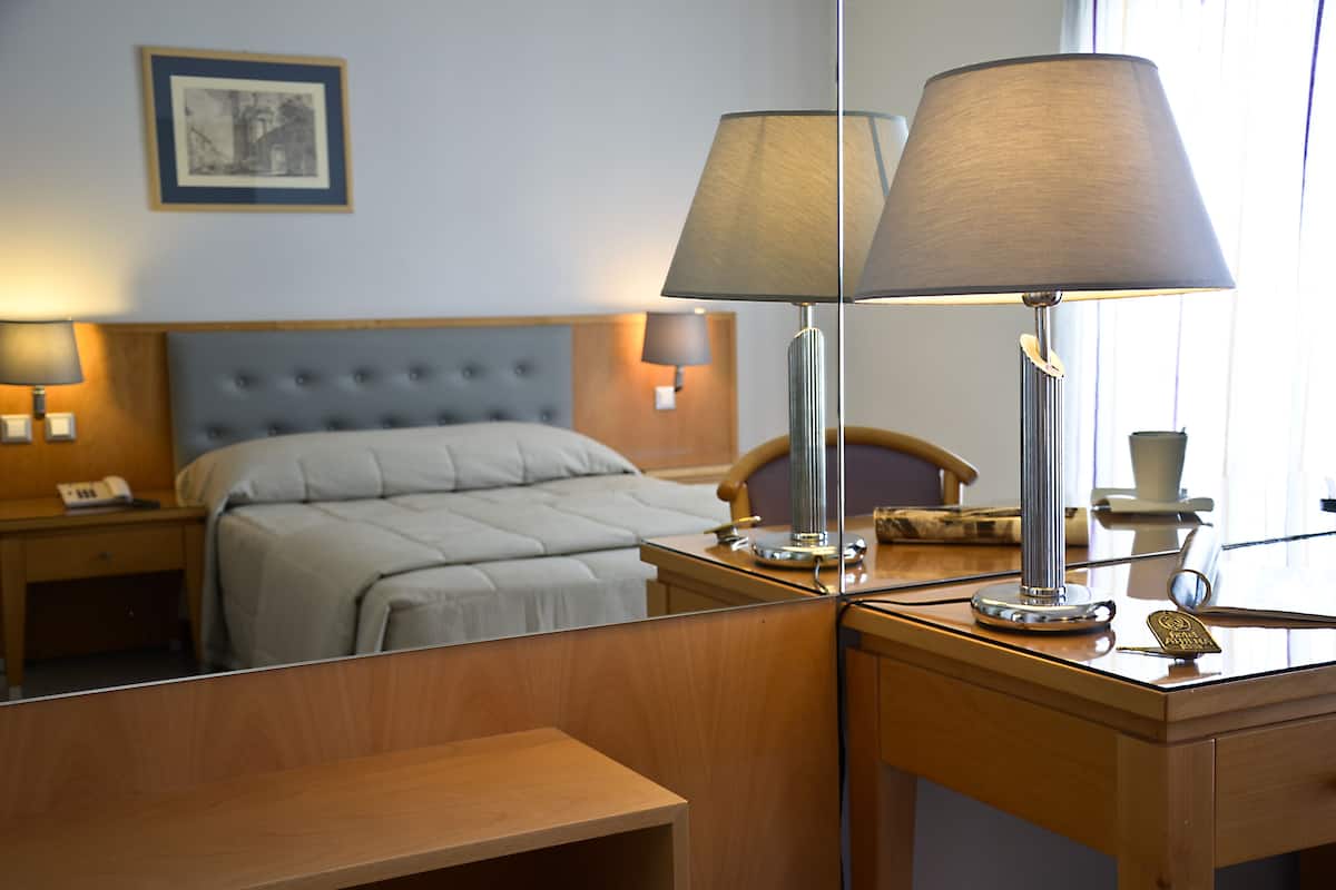 Une chambre d’hôtel avec un lit, deux lampes de chevet, un bureau avec une lampe, un téléphone et un tableau encadré au mur.