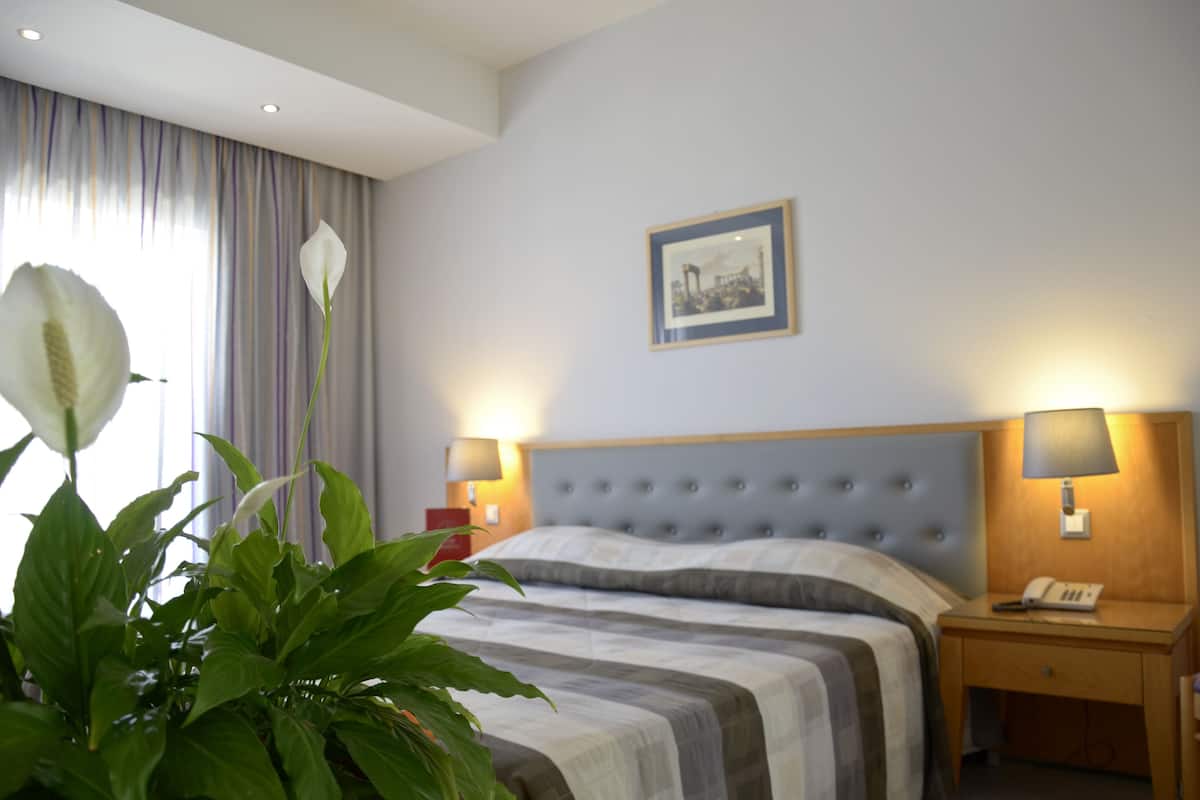 Une chambre d’hôtel avec un lit, deux lampes de chevet, une table de nuit, un tableau encadré au mur et une plante en pot.