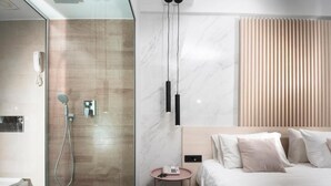 Een moderne hotelkamer met een douchecabine van glas, een nachtkastje met een telefoon, en een bed met een gewatteerde spreidekker.