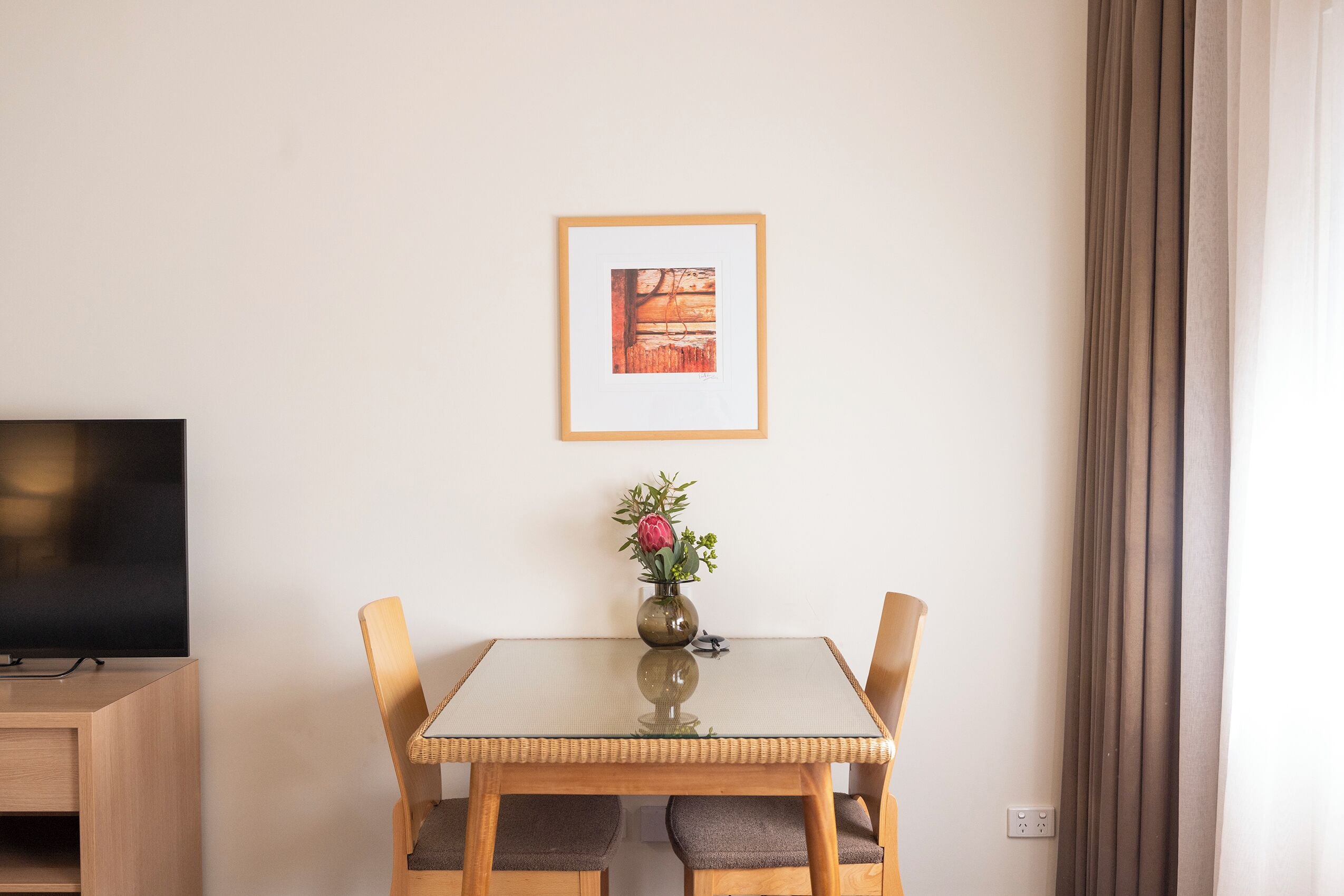 En matplats med ett glasbord, trästolar, en vas med blommor, en inramad tavla på väggen och en tv-apparat på ett träskåp.