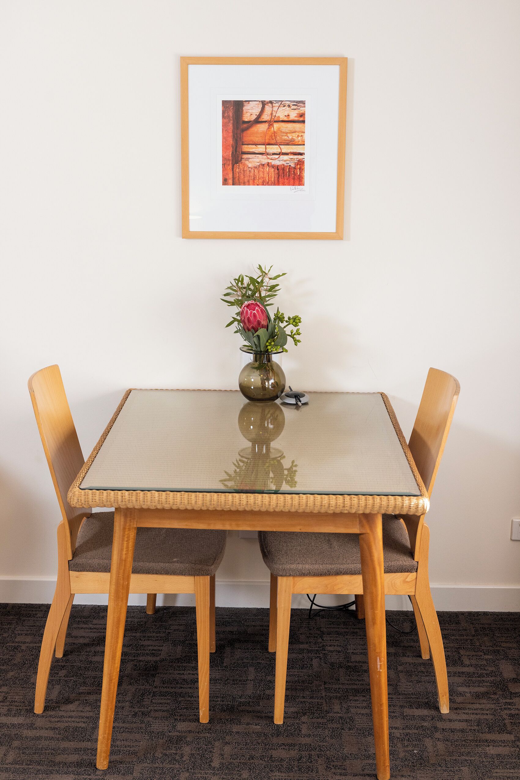 A dining table with a glass top and four wooden chairs, a vase with flowers, and a framed picture on the wall.