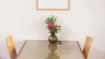A dining table with a glass top and four wooden chairs, a vase with flowers, and a framed picture on the wall.
